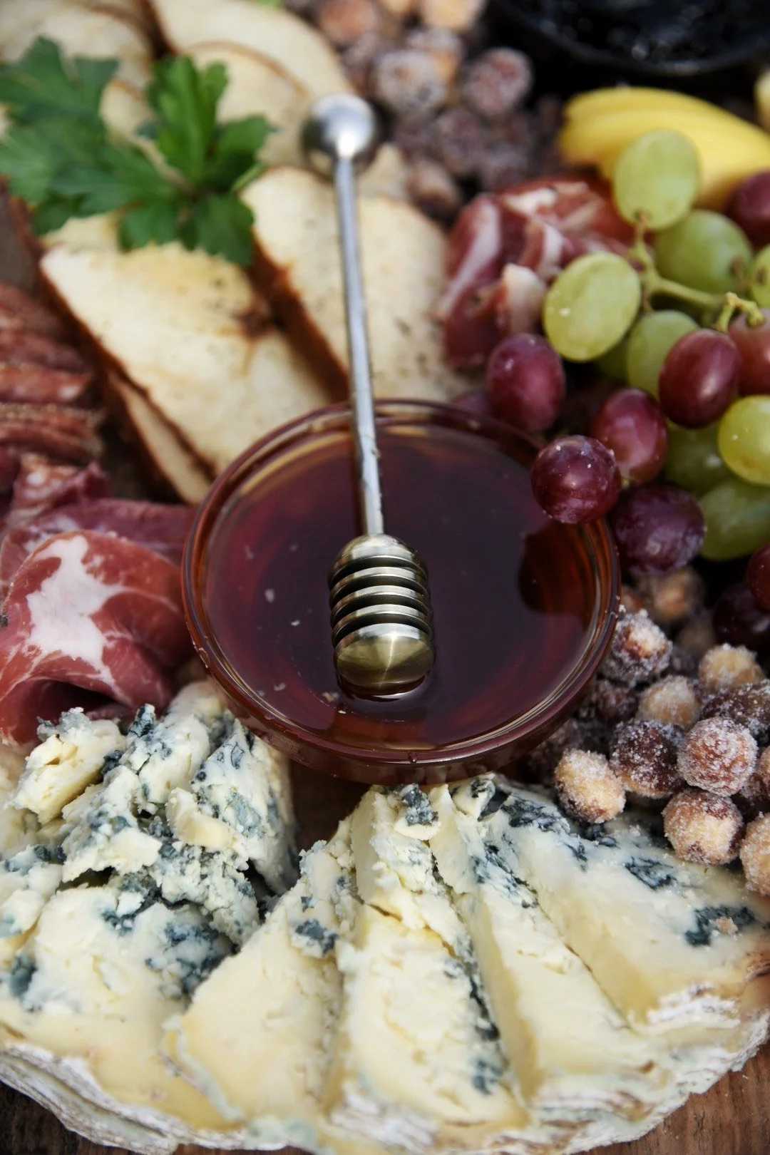 A cheese platter with blue cheese, brie, sliced cured meats, crackers, green and red grapes, and a small bowl of honey with a honey dipper.