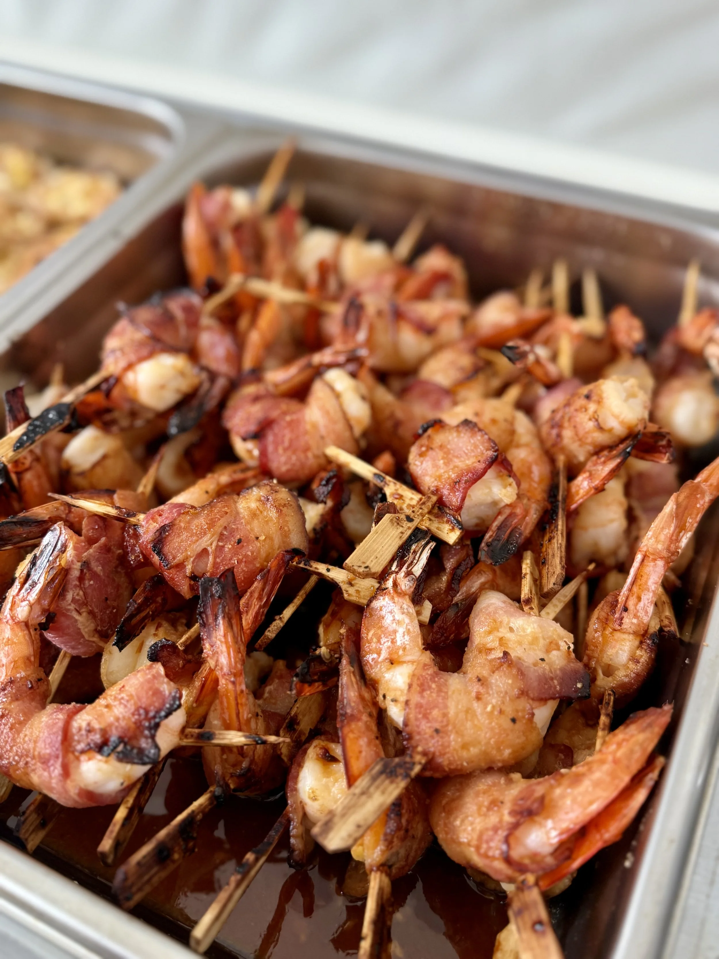 Close-up of grilled shrimp wrapped in bacon on skewers, placed in a metal tray.