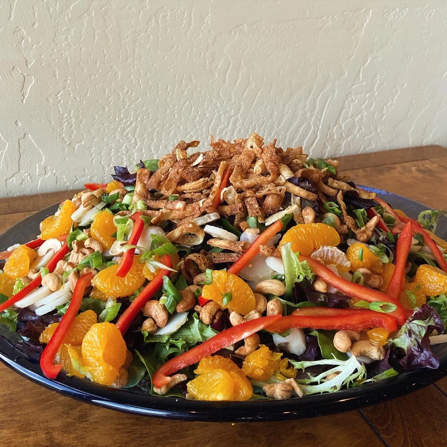 A colorful salad on a black plate featuring mixed greens, orange slices, red bell pepper strips, chopped green onions, cashews, and crispy fried onion topping.
