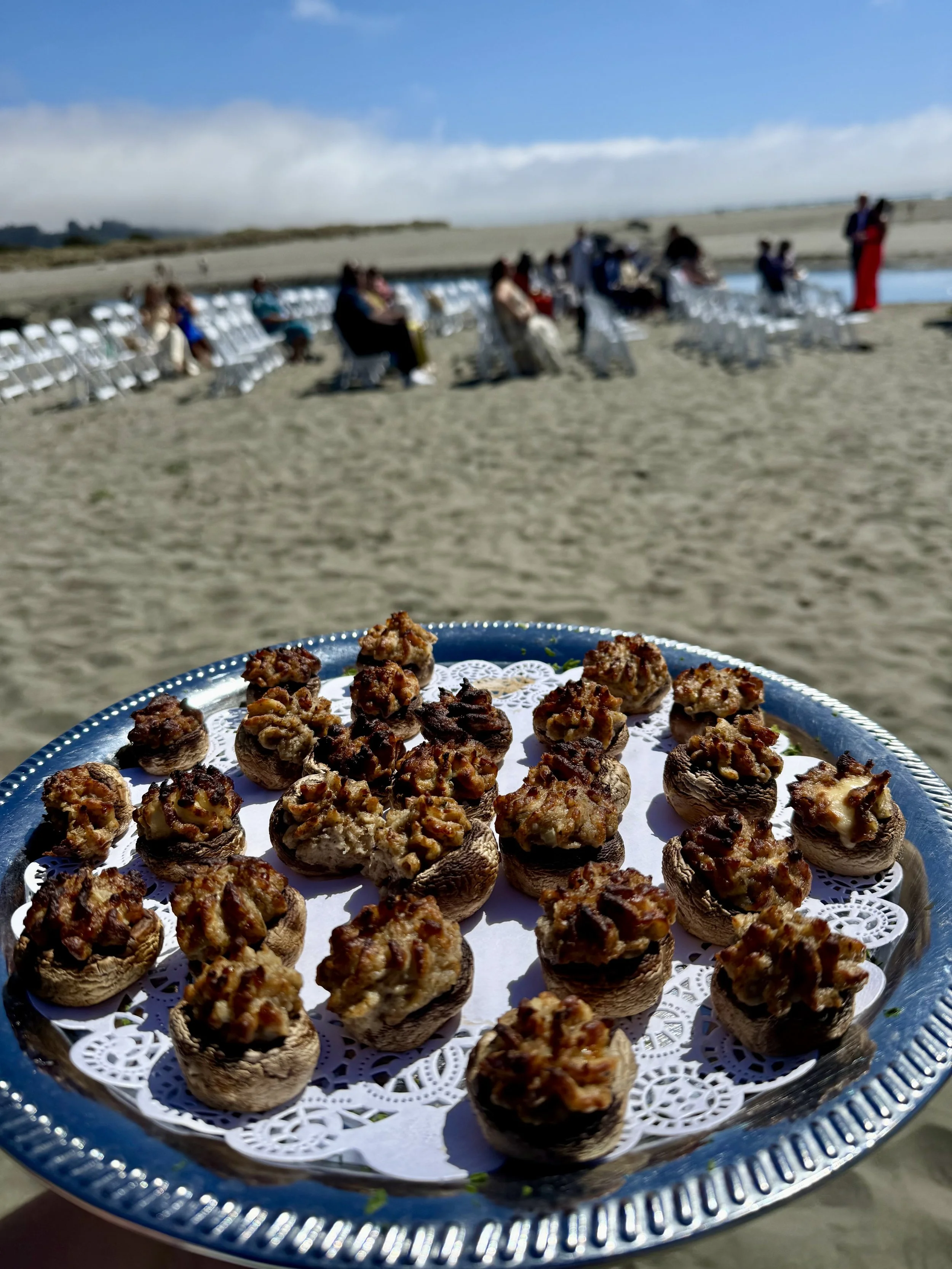 Tray of stuffed mushrooms with meat topping held against a beach backdrop with people sitting and standing near a body of water