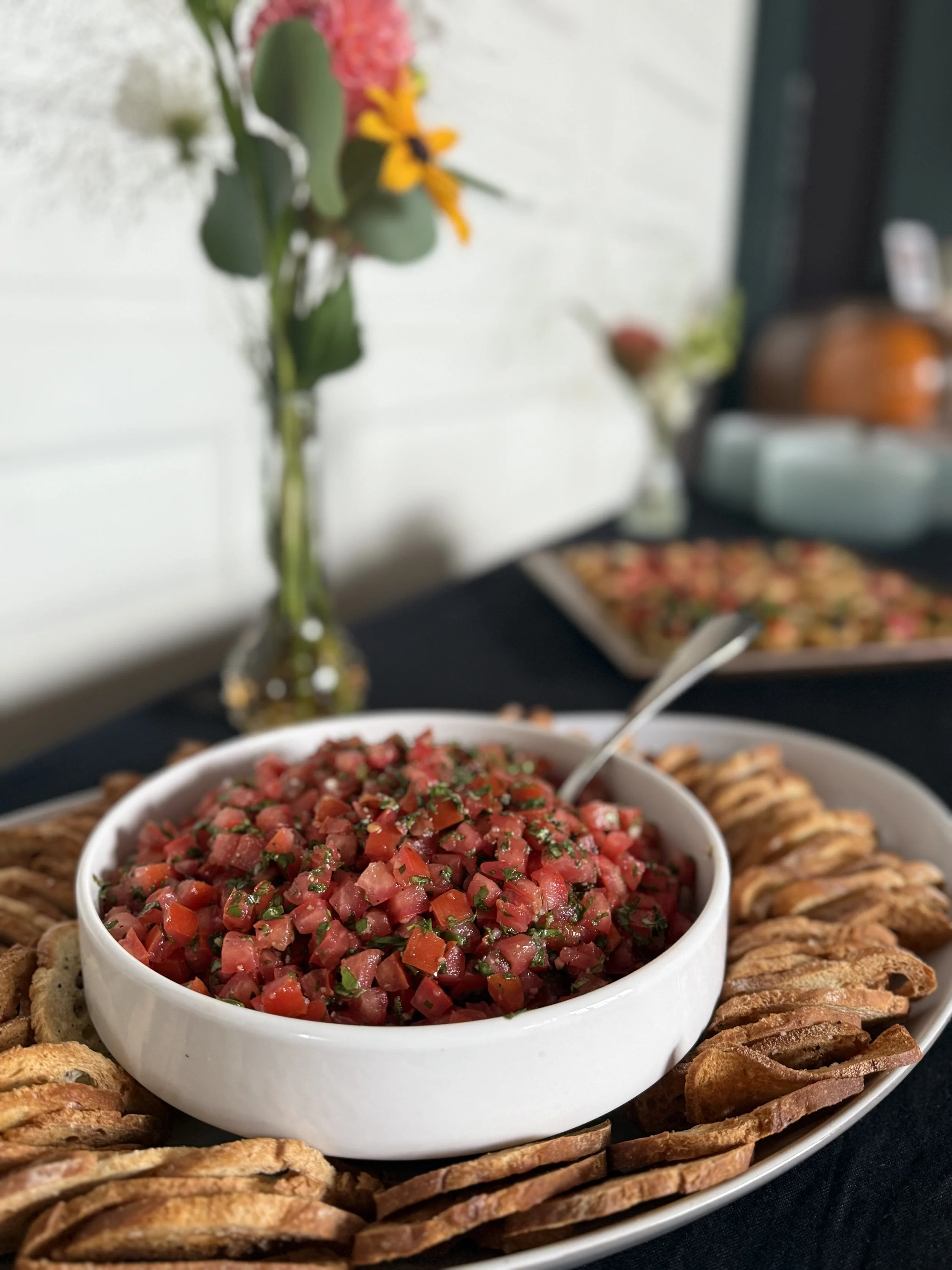 A white bowl of pico de gallo surrounded by toasted flour tortillas on a black table, with a vase of flowers in the background.