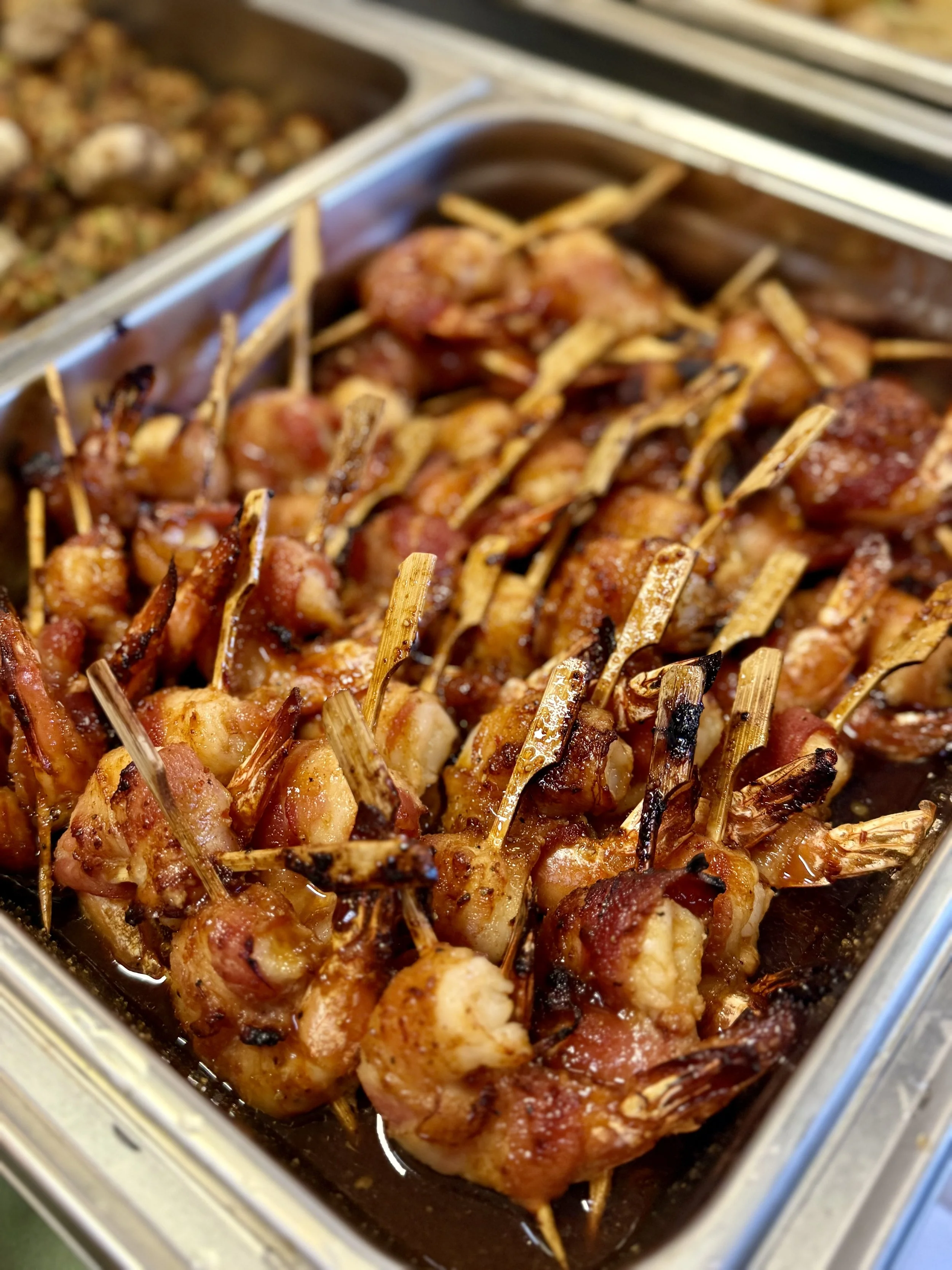 Tray of cooked bacon-wrapped shrimp skewers in a buffet setting.