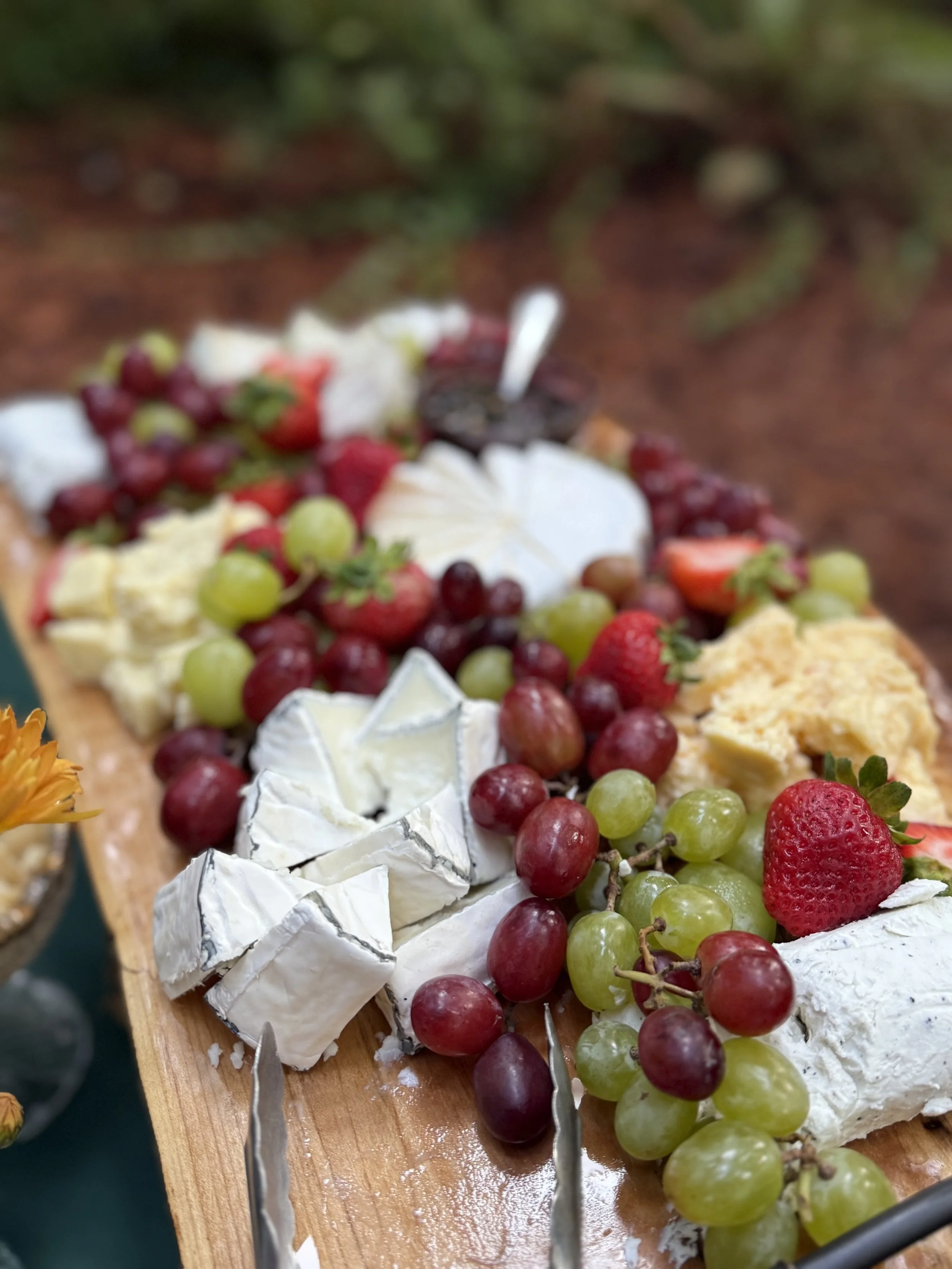 A cheese platter with various cheeses, grapes, and strawberries on a wooden board outdoors.