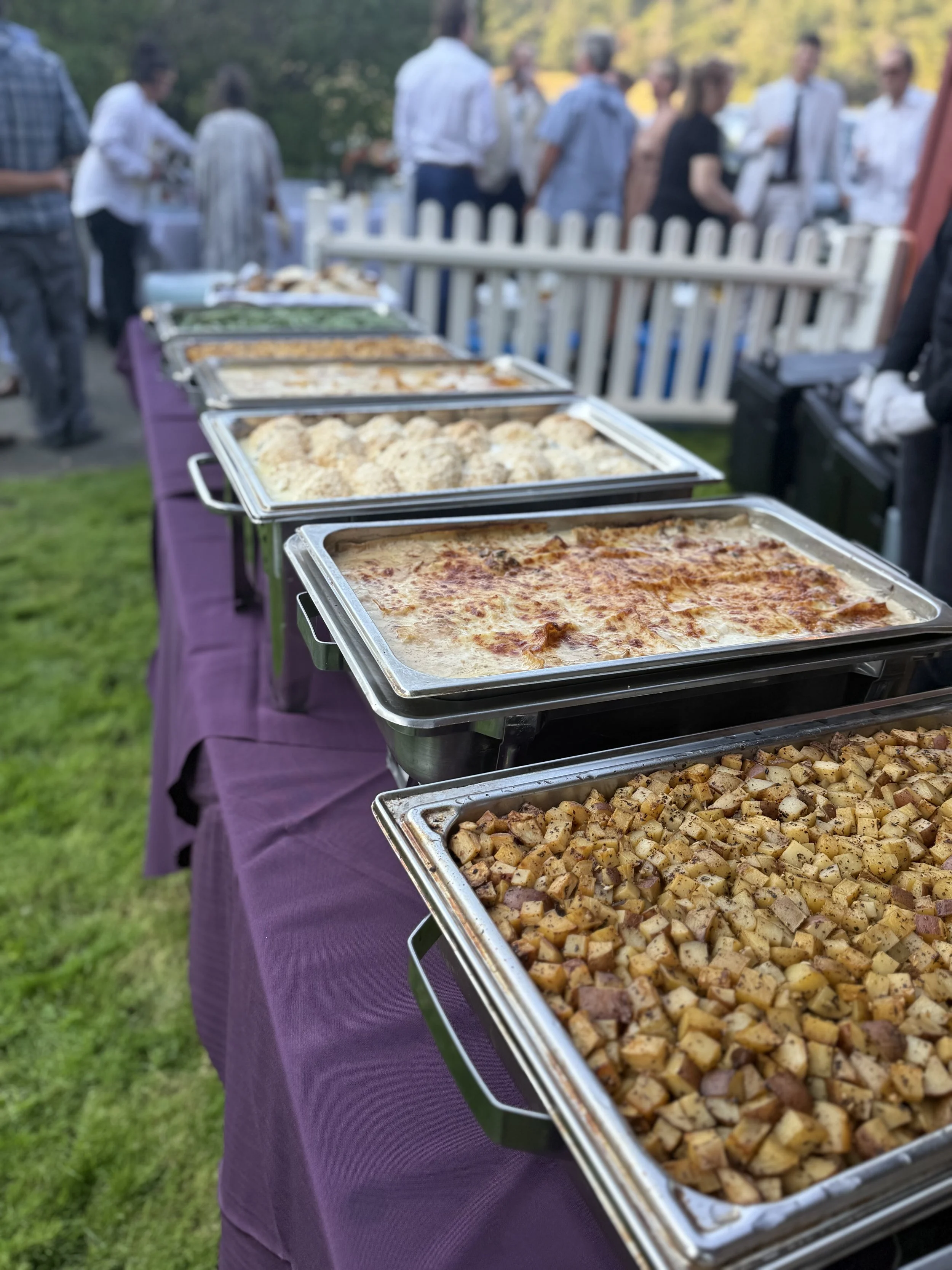 Buffet table with trays of baked dishes, including diced potatoes, lasagna, and bread rolls, at an outdoor gathering with people in the background.