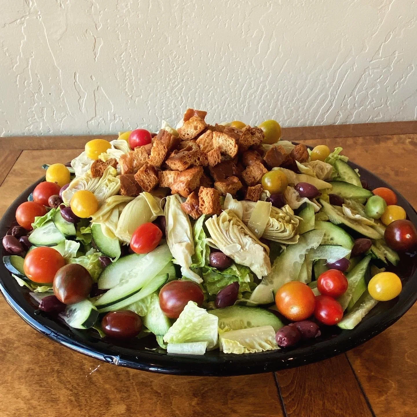 Large bowl with mixed salad including cherry tomatoes, cucumber slices, lettuce, artichoke hearts, and croutons on top, placed on a wooden table against a textured wall.