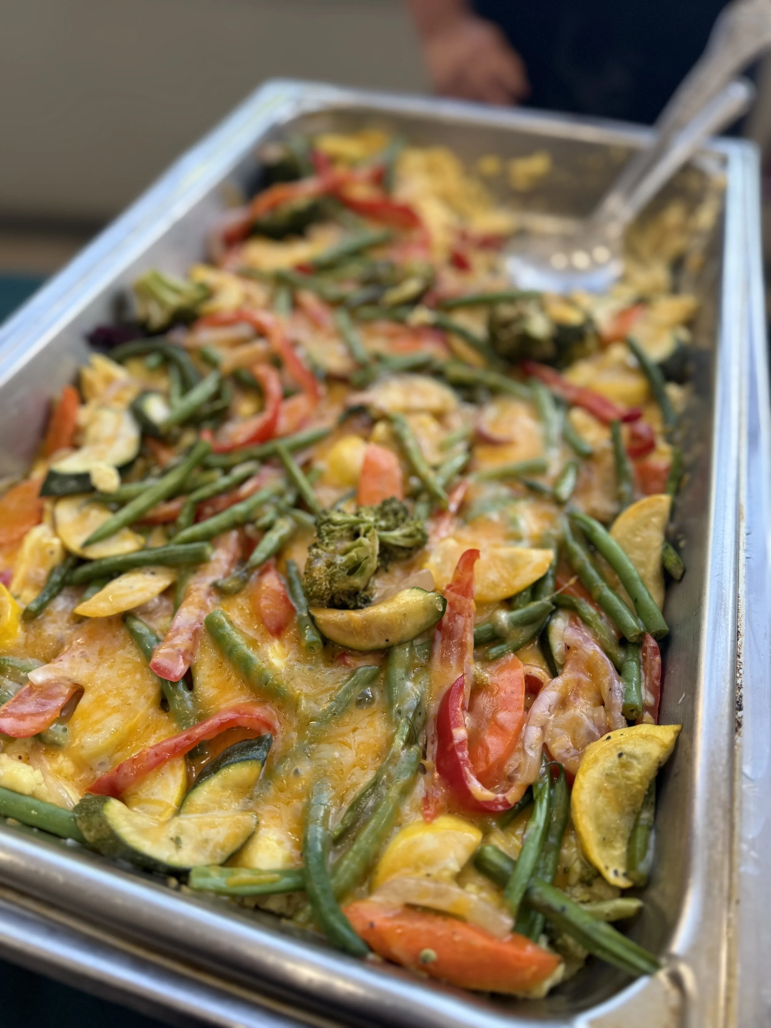 Colorful casserole dish with green beans, sliced bell peppers, broccoli, and cheese in a metal serving tray.