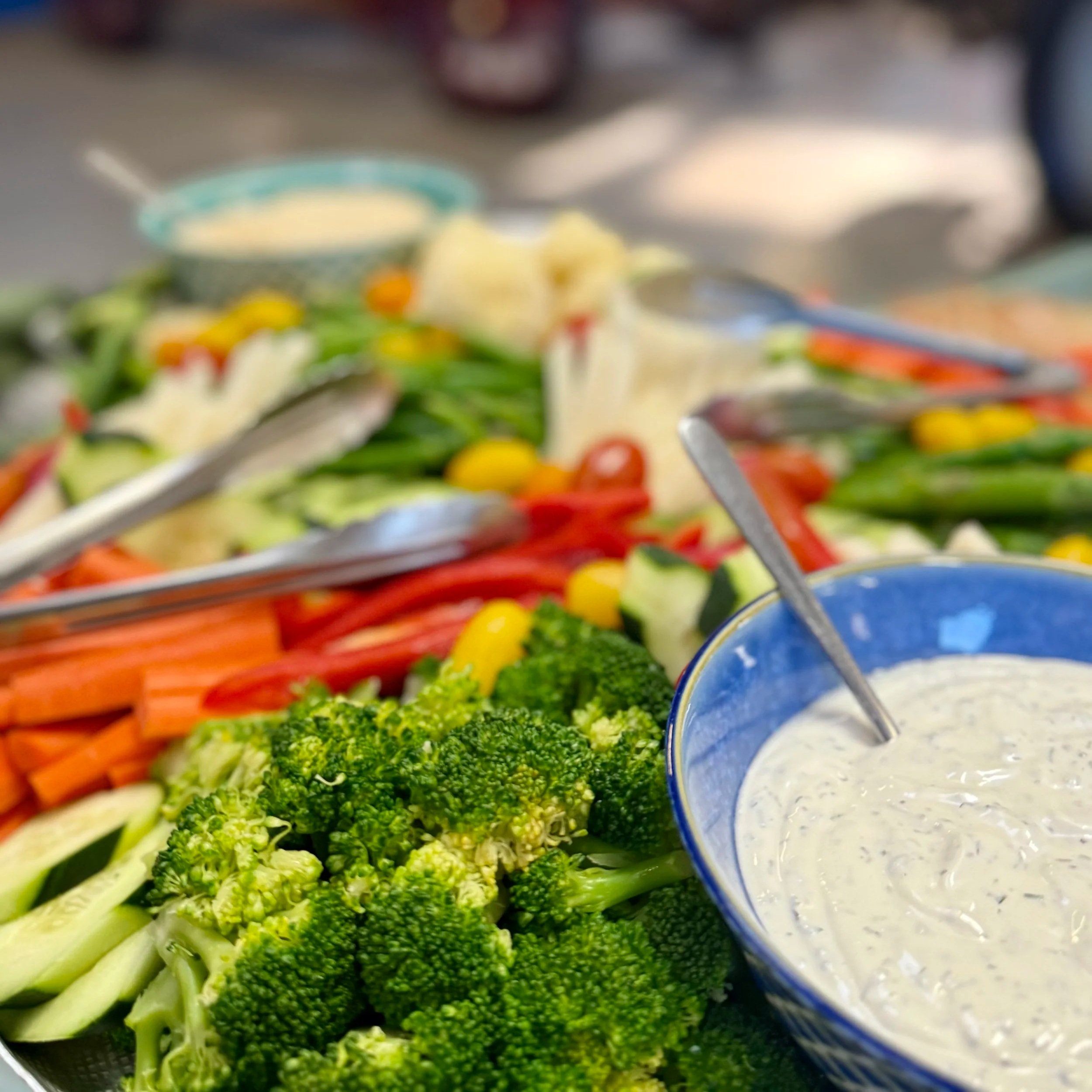 Colorful fresh vegetables including broccoli, cherry tomatoes, carrots, cucumbers, and yellow peppers, with bowls of dip or dressing, on a serving table.