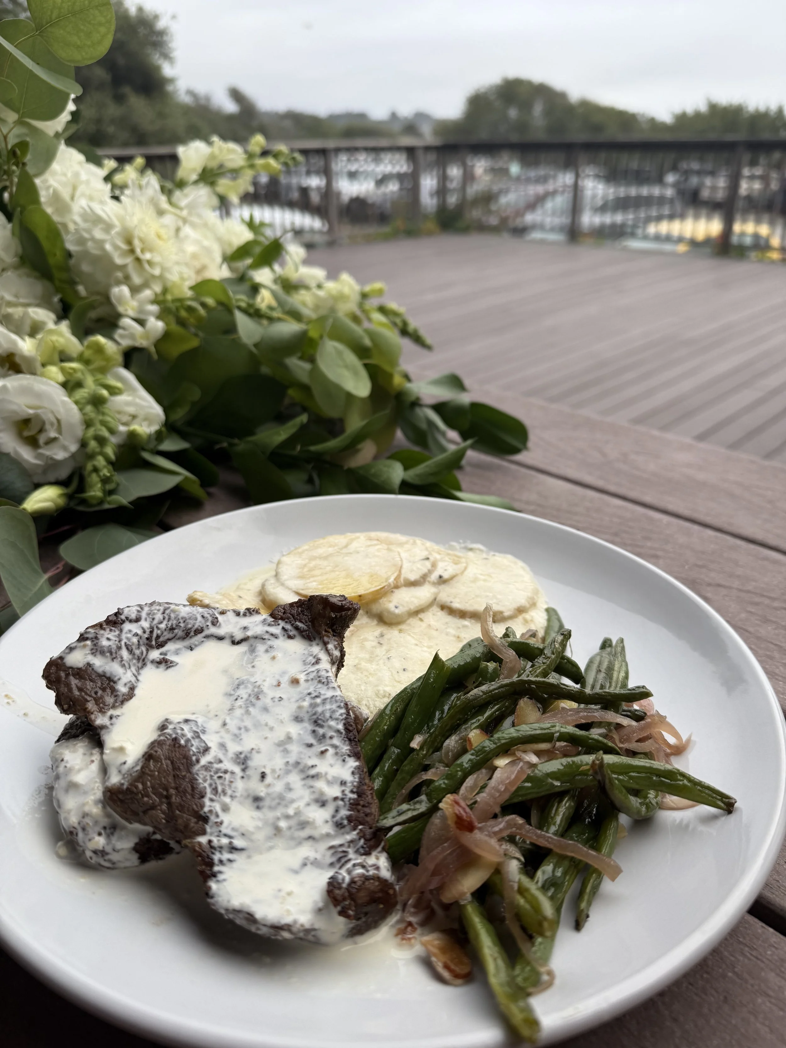 A plate of food with a piece of meat covered in cream sauce, a serving of sliced potatoes, and green beans with onions. The plate is on a wooden table outside, with a bouquet of white flowers nearby and a blurred outdoor railing in the background.