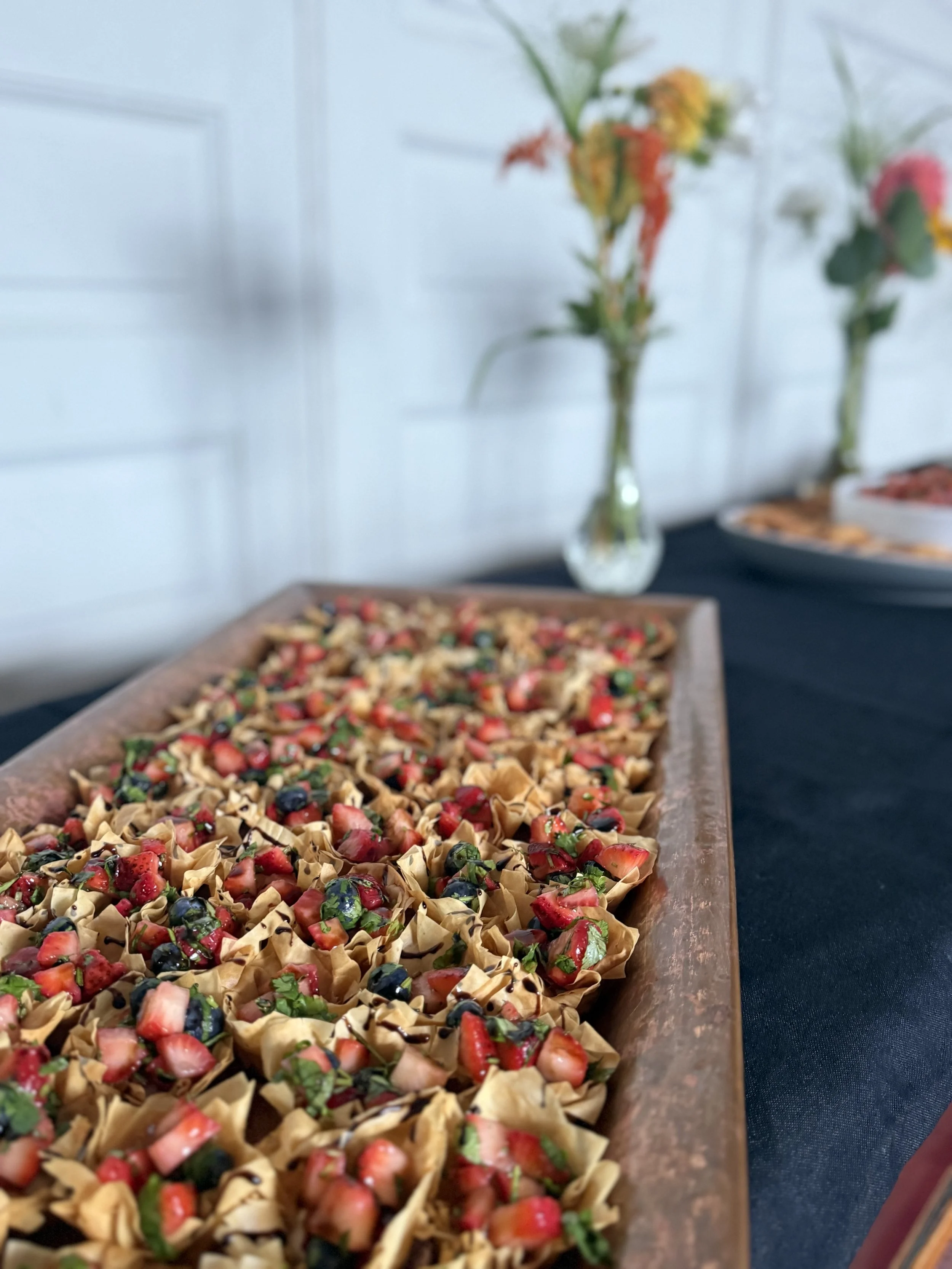 A long rectangular serving platter filled with bite-sized dessert cups topped with strawberries, blueberries, and chopped herbs, on a black tablecloth with a blurred background of vases with flowers and plates of food.