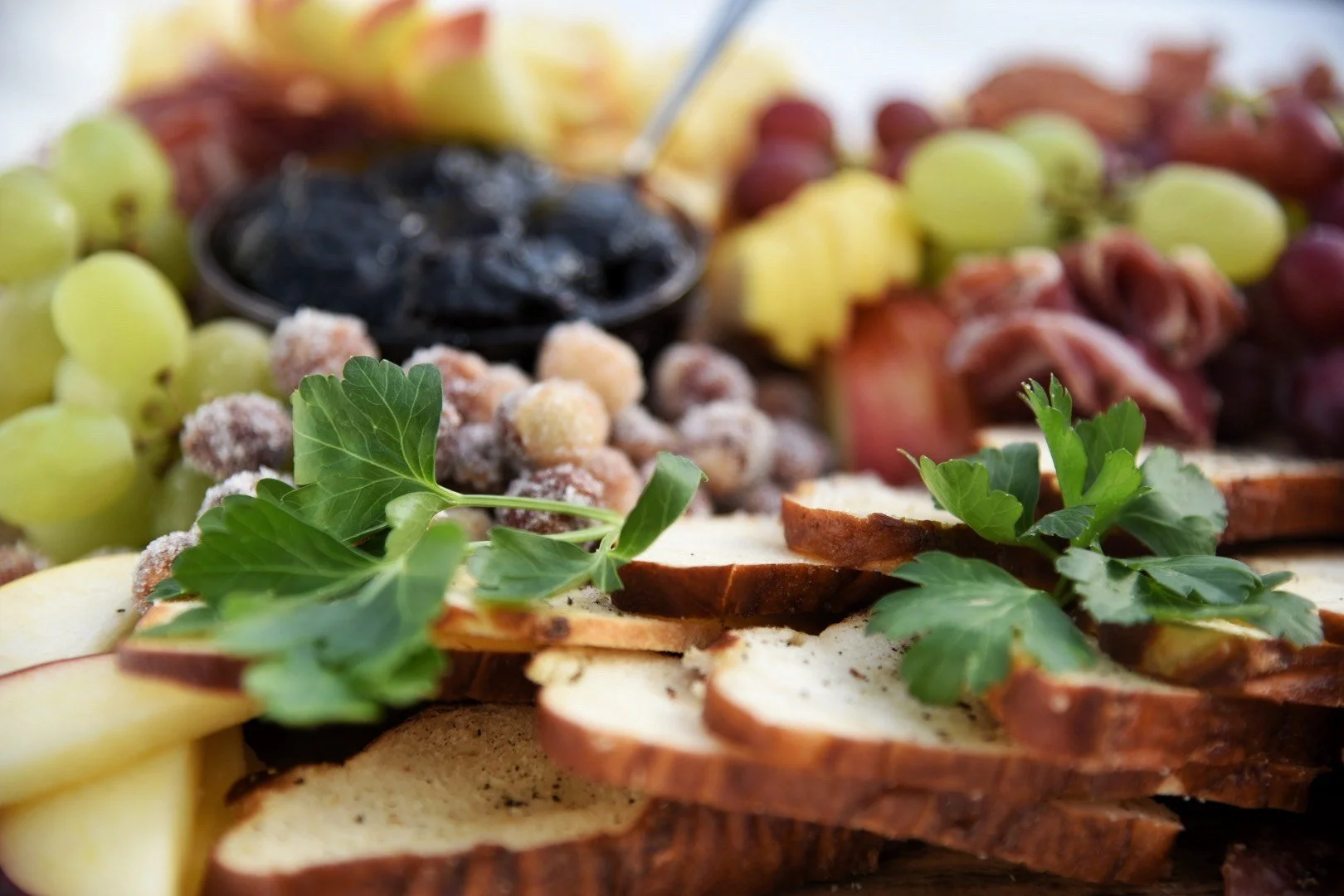Close-up of a charcuterie board with grapes, sliced bread, and garnishes including parsley, with cheese, dried fruit, and dips in the background.