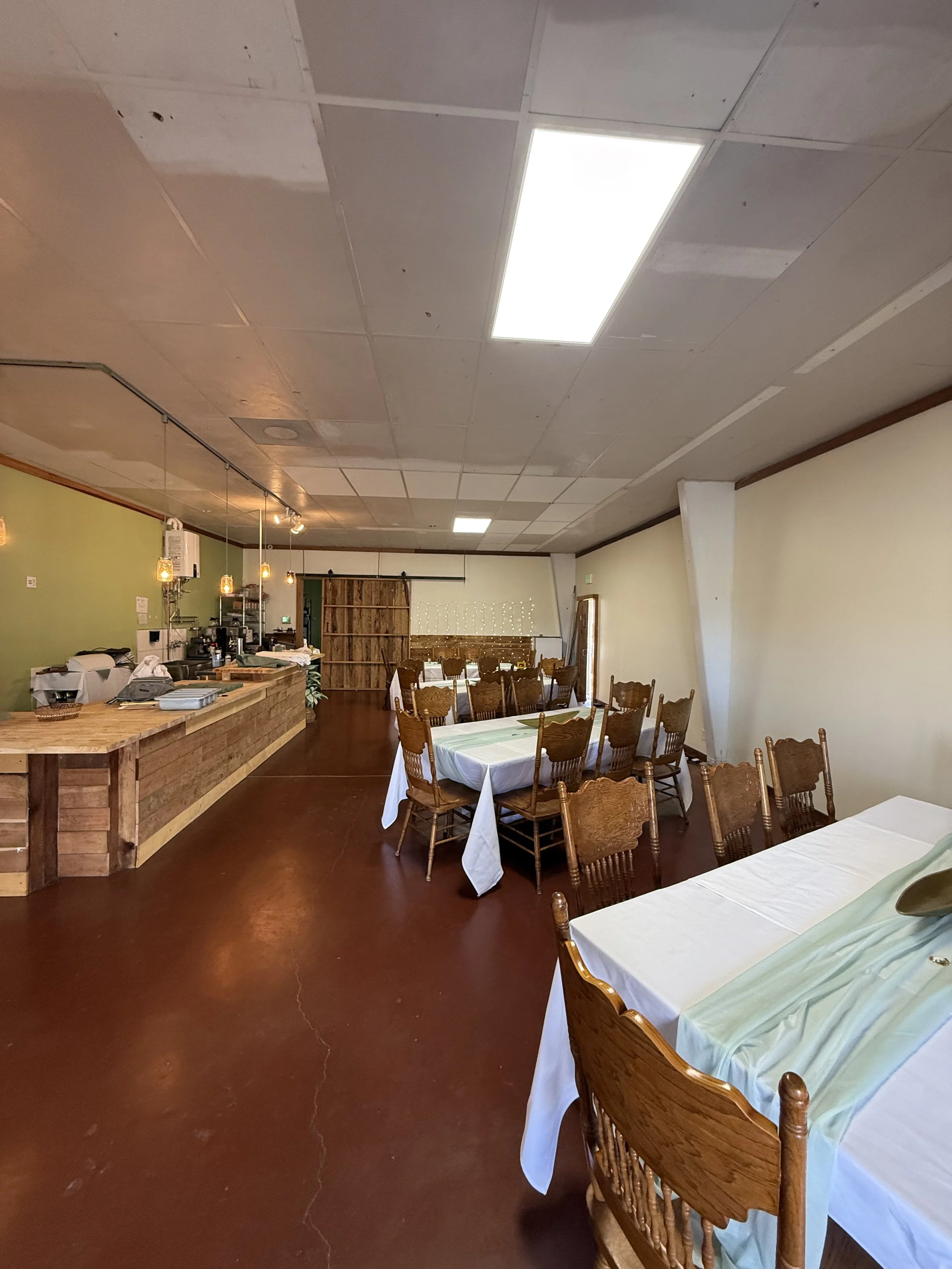 Empty restaurant dining area with wooden tables and chairs, some tables have white tablecloths, and a wooden counter in the background. Indoor lighting and a green accent wall.