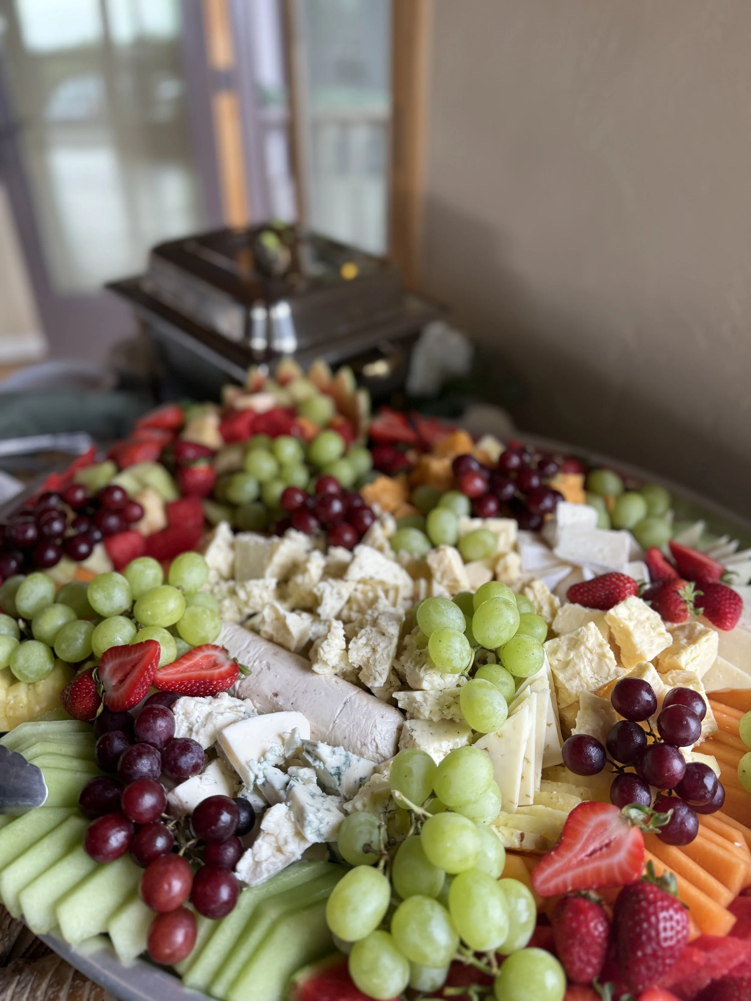 A cheese and fruit platter with grapes, strawberries, honeydew, cantaloupe, carrots, and various cheeses, with a chafing dish in the background.