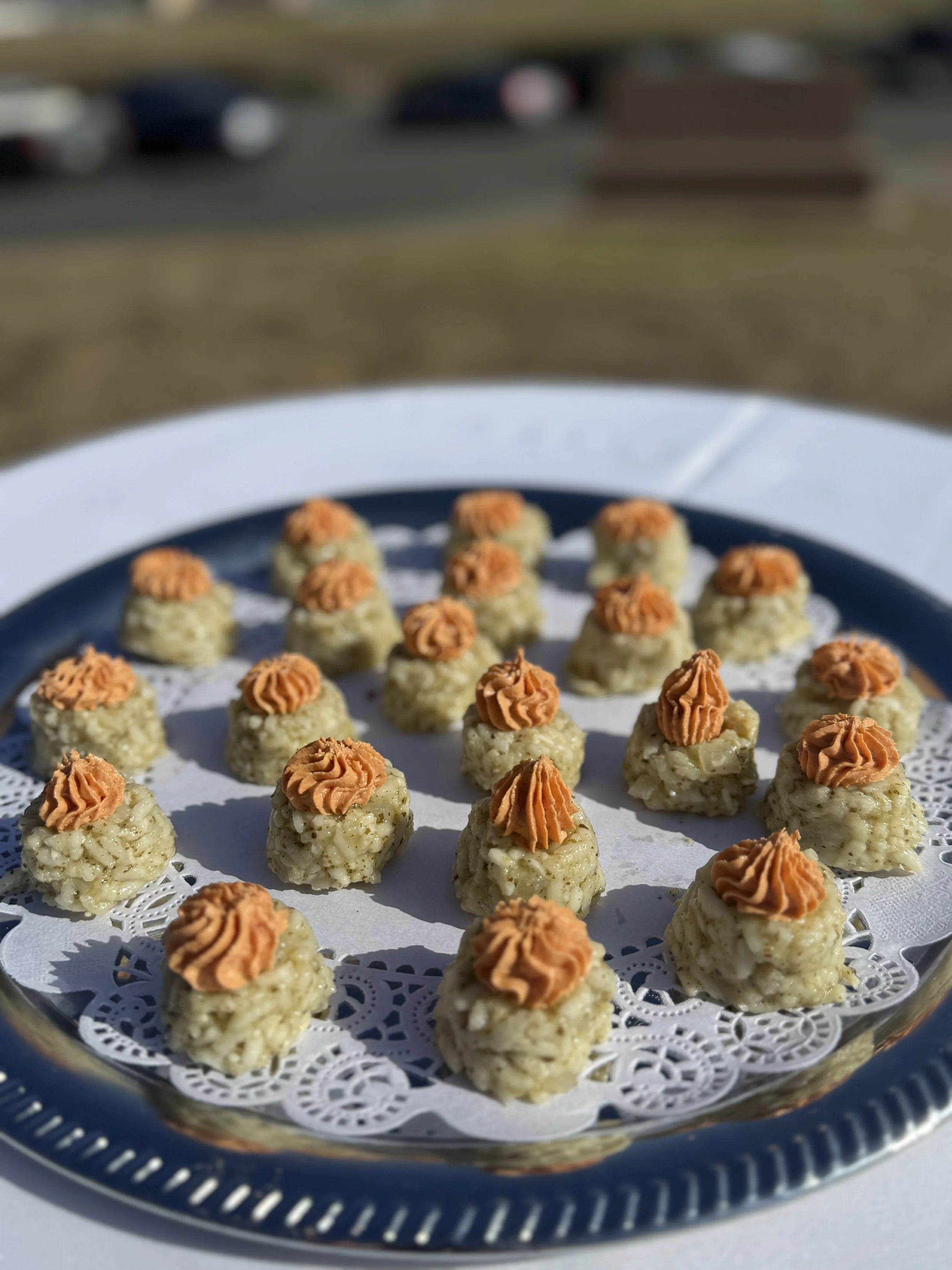 Close-up of bite-sized appetizers with a dollop of orange sauce on top, arranged on a silver tray with a paper doily, outdoors on a sunny day.