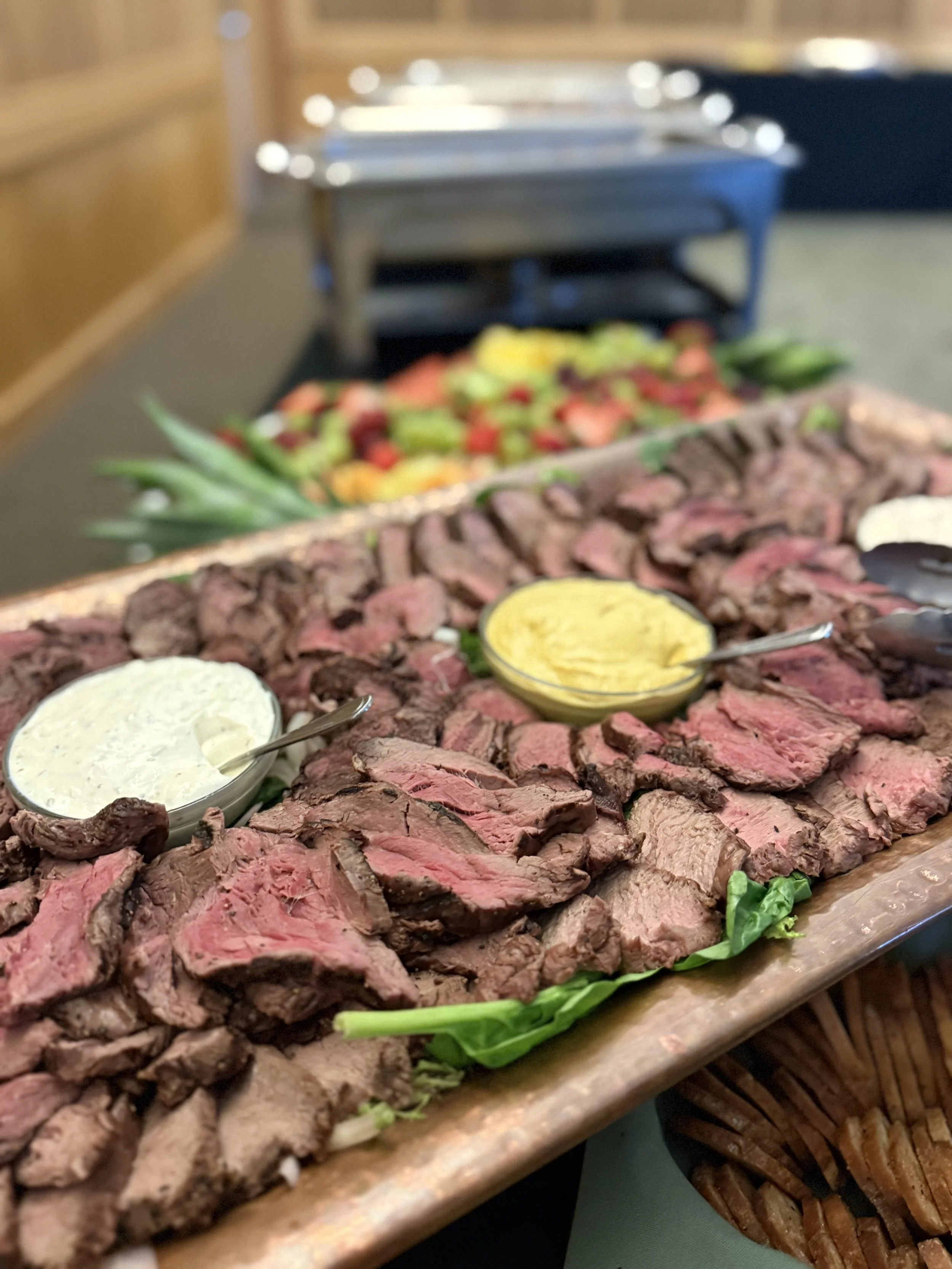 Sliced cooked beef with creamy sauces on a wooden platter. In the background, a fruit salad with strawberries, pineapple, and other fruits.