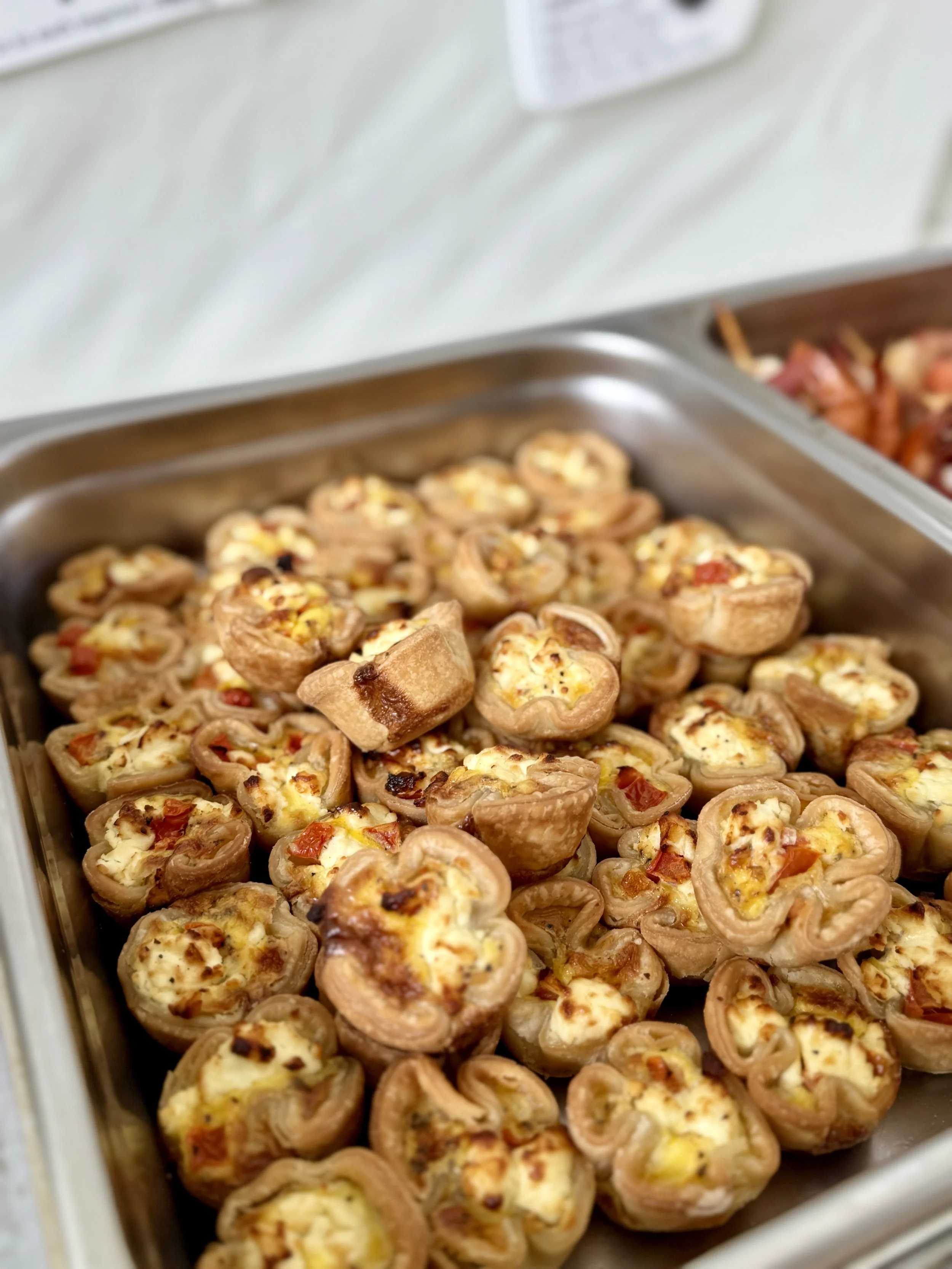Miniature savory tarts filled with cheese, diced tomatoes, and possibly eggs, arranged on a metal tray at a buffet or catering event.