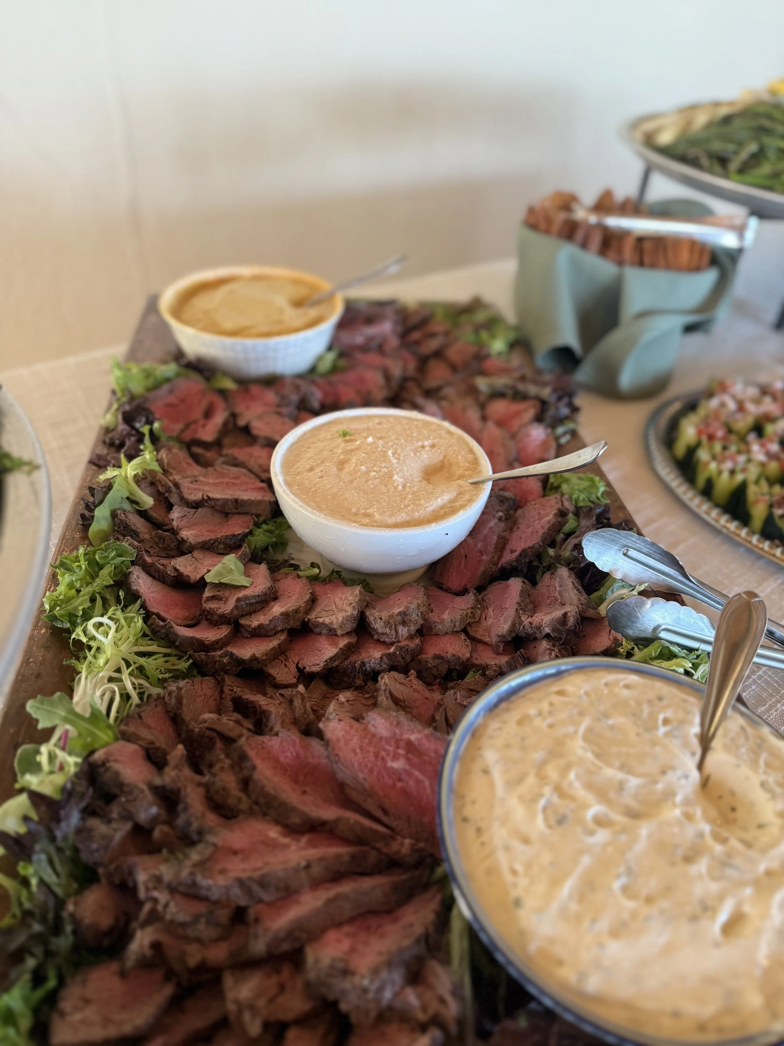 A platter of sliced beef with three bowls of different sauces or dressings around it, and additional dishes in the background.