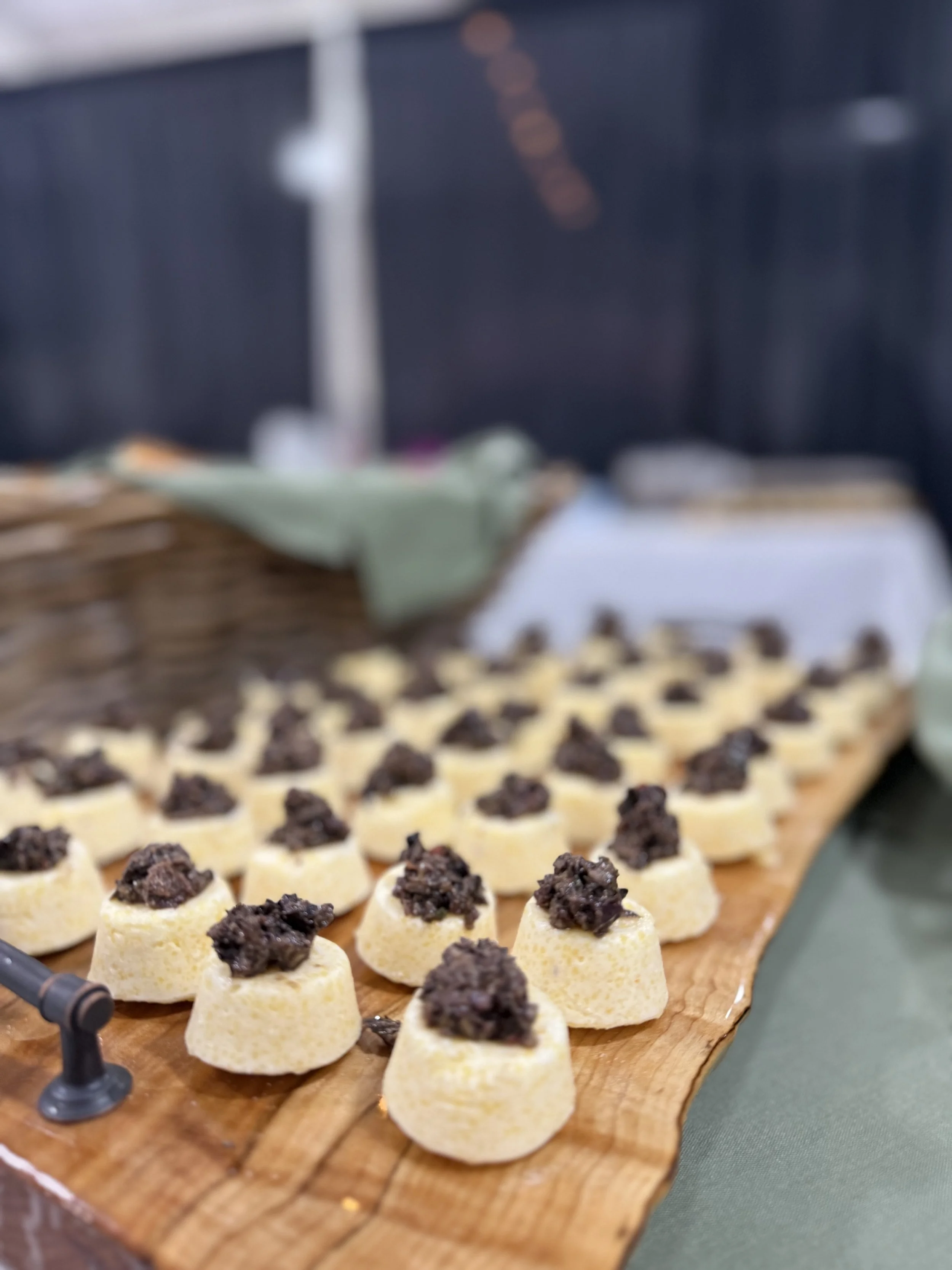 Close-up of small, round appetizers with a topping of dark, minced meat on a wooden platter at a buffet or food event.