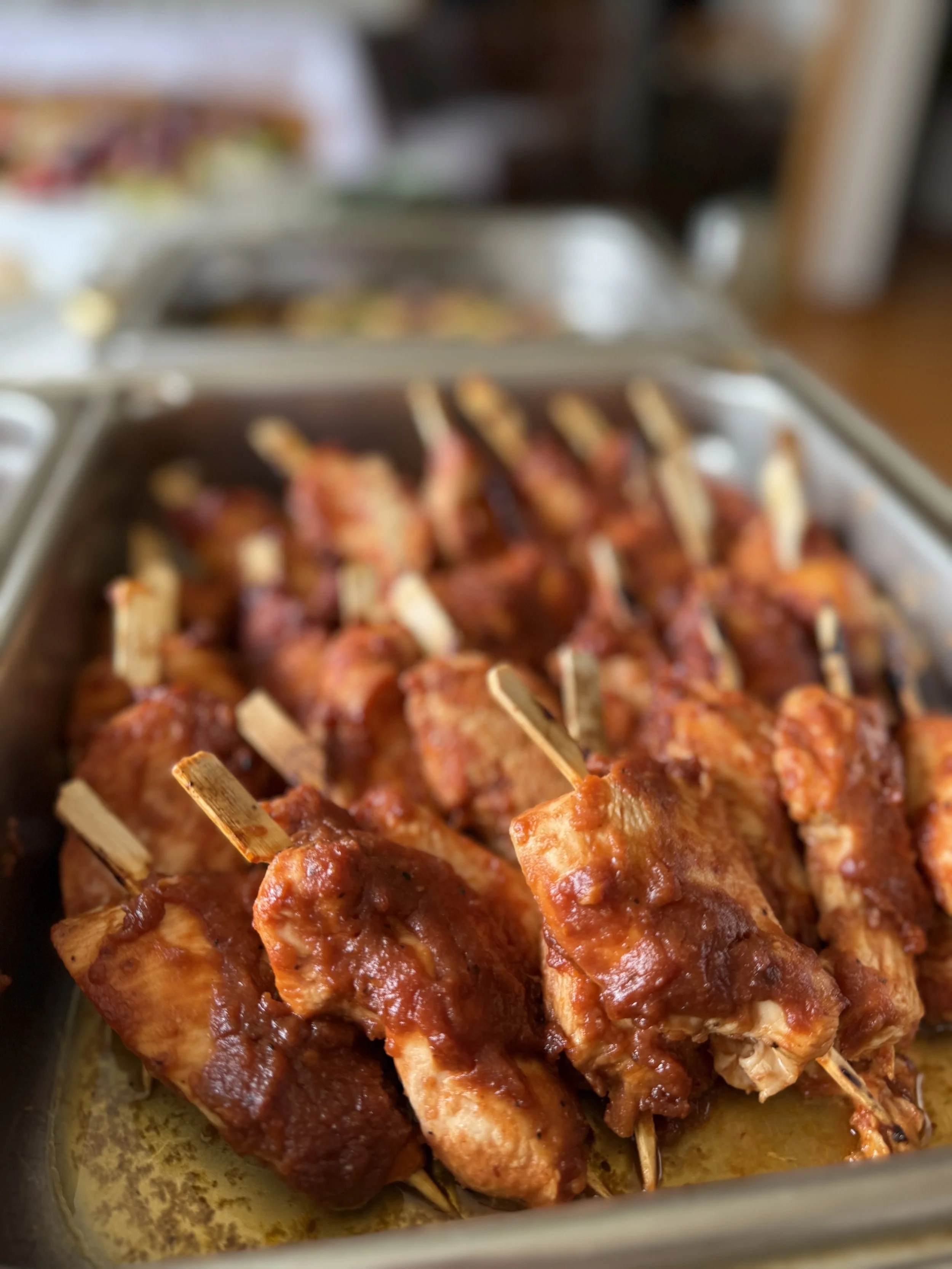 Close-up of skewered barbeque chicken drumsticks covered in sauce in a metal tray.