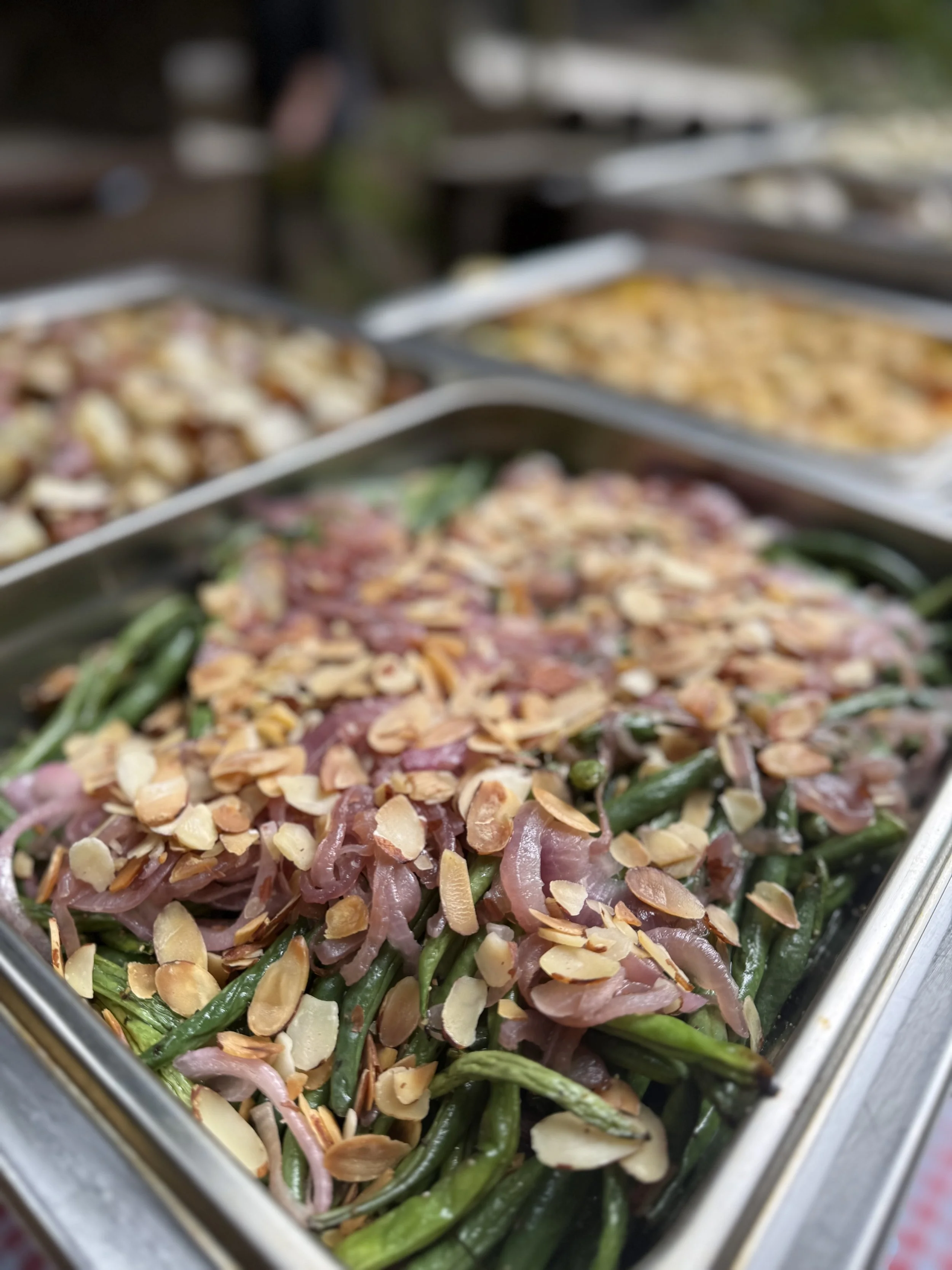 Close-up of a tray of cooked green beans topped with sliced almonds and caramelized onions at a buffet or food service setting.