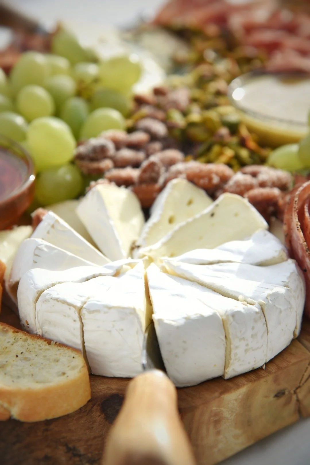 Close-up of a cheese platter including a wheel of Brie cheese cut into wedges, green grapes, nuts, and sliced bread on a wooden serving board.
