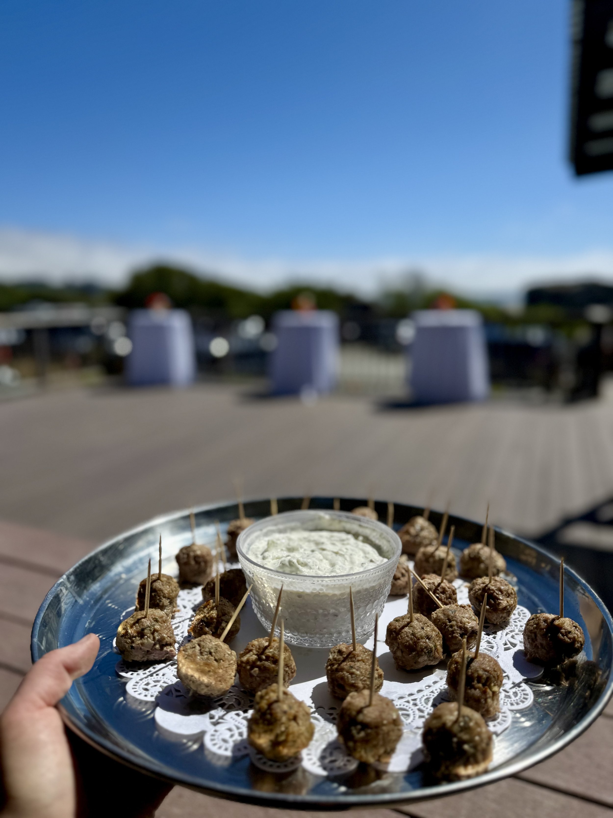 Tray of meatballs with toothpicks and a bowl of creamy dip held by a hand outdoors with a clear blue sky.