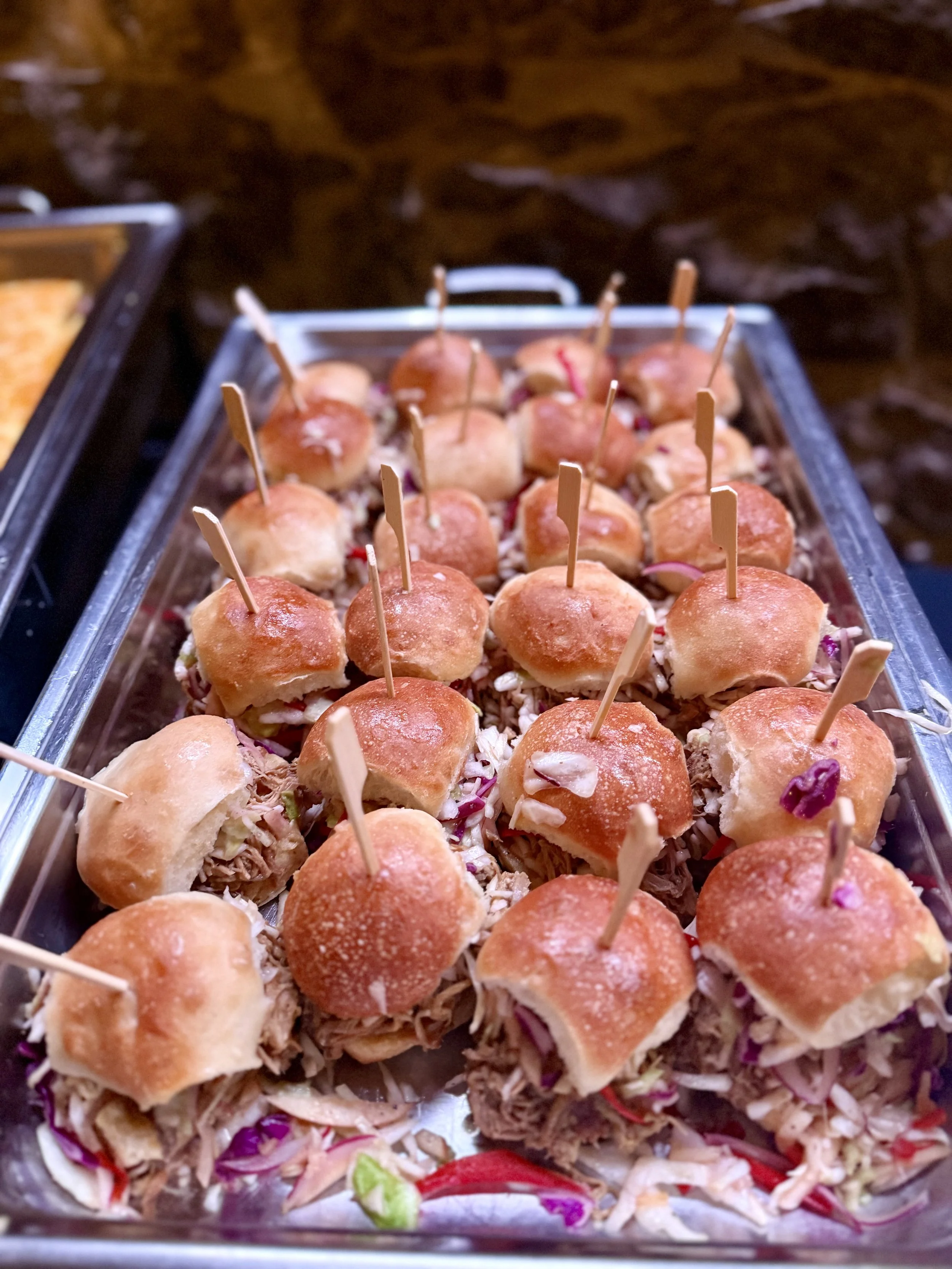 Tray of small sandwiches with slider buns filled with shredded meat, topped with pickled red onions, served with toothpicks.
