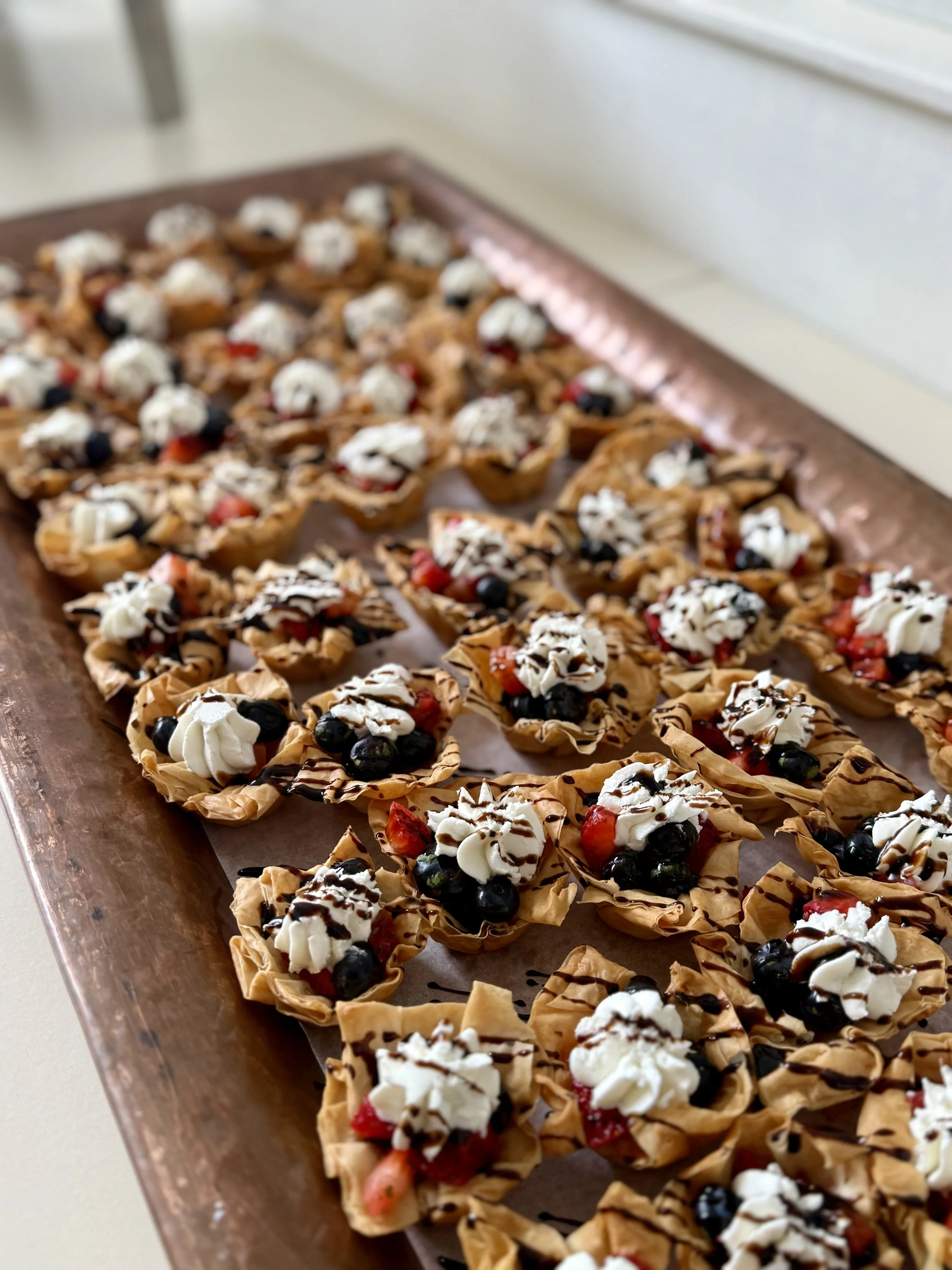 Tray of small pastry bites topped with whipped cream, blueberries, strawberries, and drizzled with chocolate.