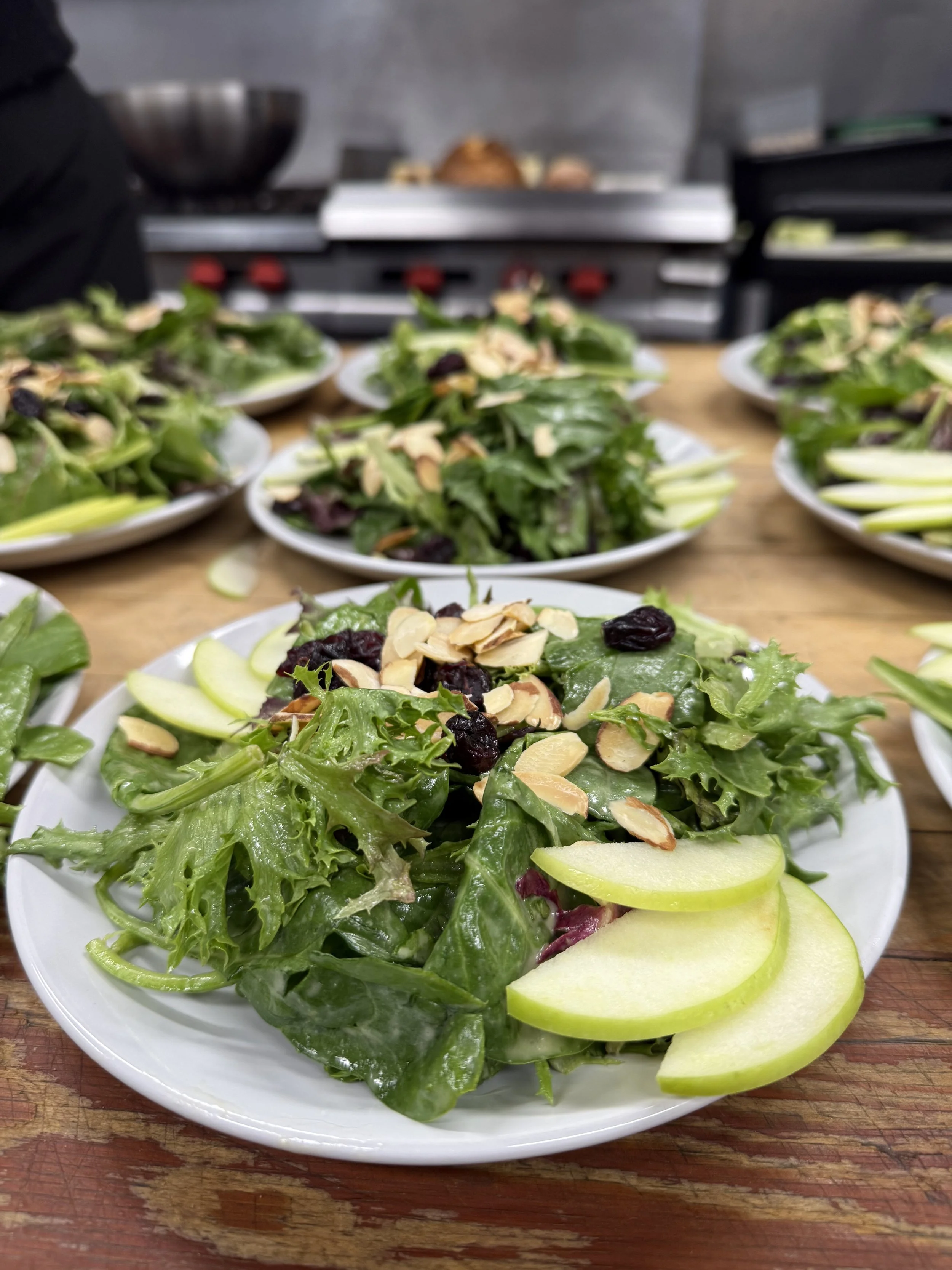 Salad plates with mixed greens, sliced green apples, dried cranberries, and almond slices on a wooden surface in a kitchen setting.