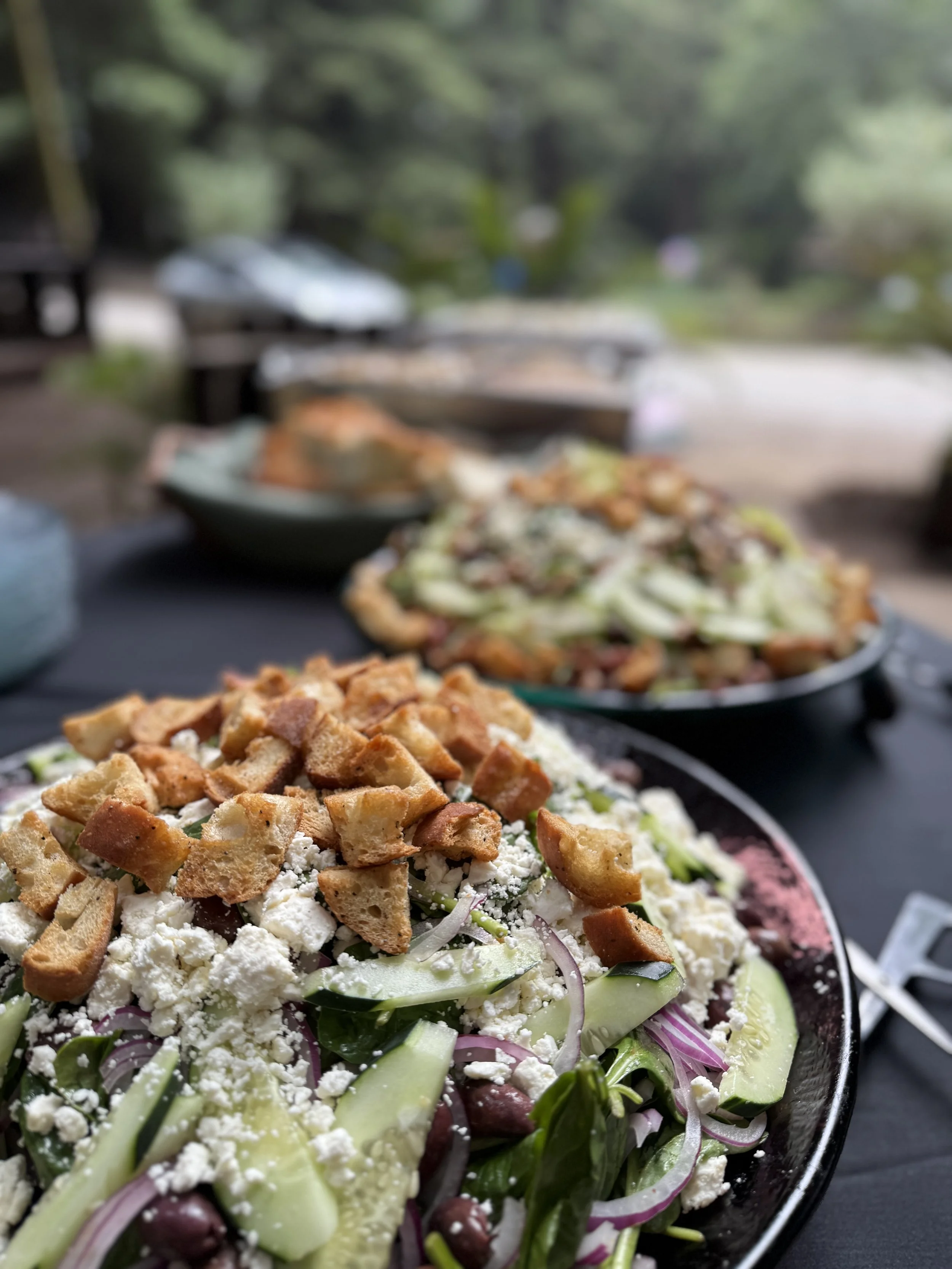 Close-up of a fresh salad topped with chopped bread, feta cheese, cucumber, red onion, and olives, set outdoors on a black table with other dishes in the background.