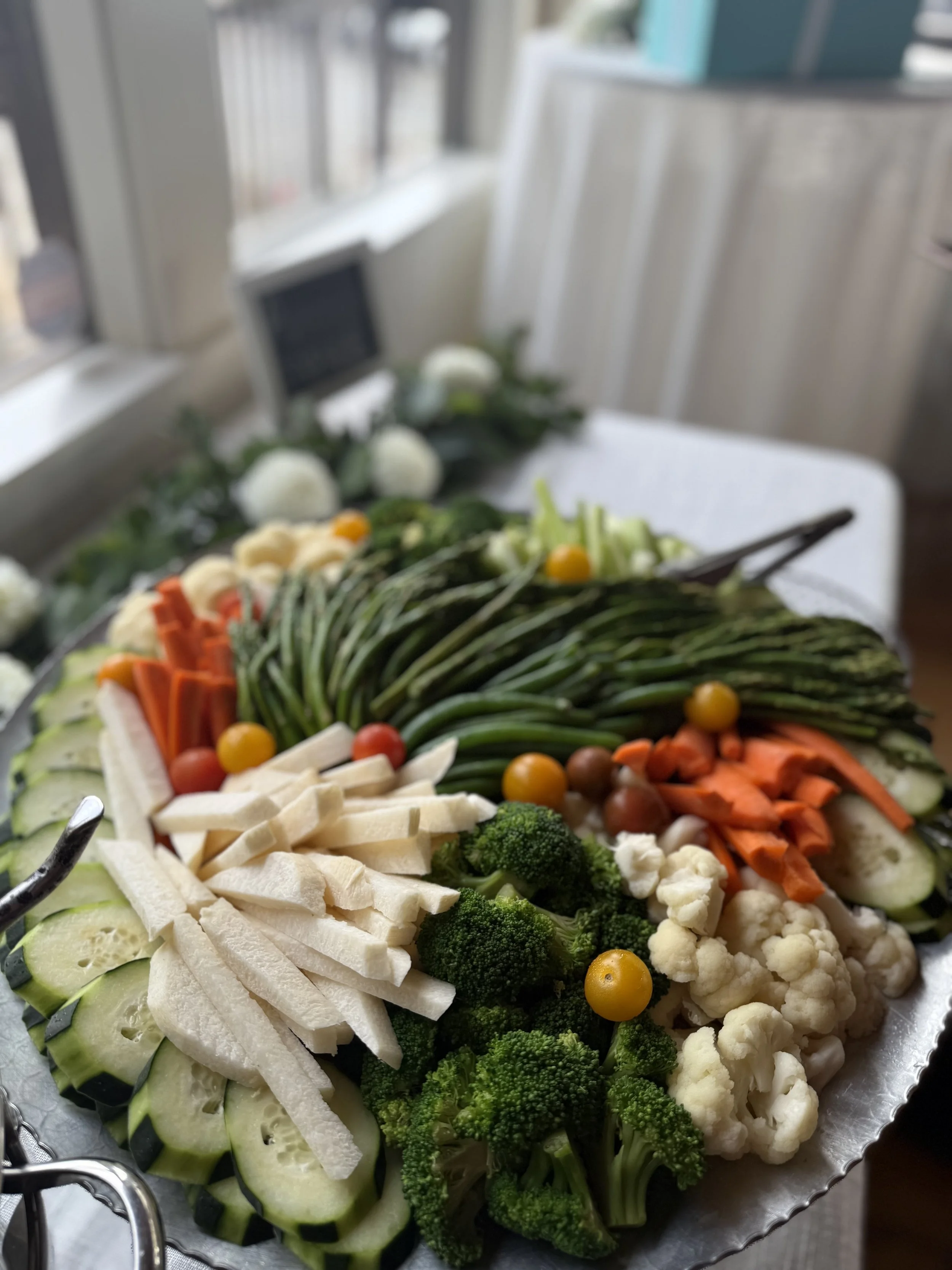 A tray of assorted fresh vegetables including cucumbers, cauliflower, broccoli, cherry tomatoes, carrots, green beans, and white cheese sticks, placed on a table near a window.