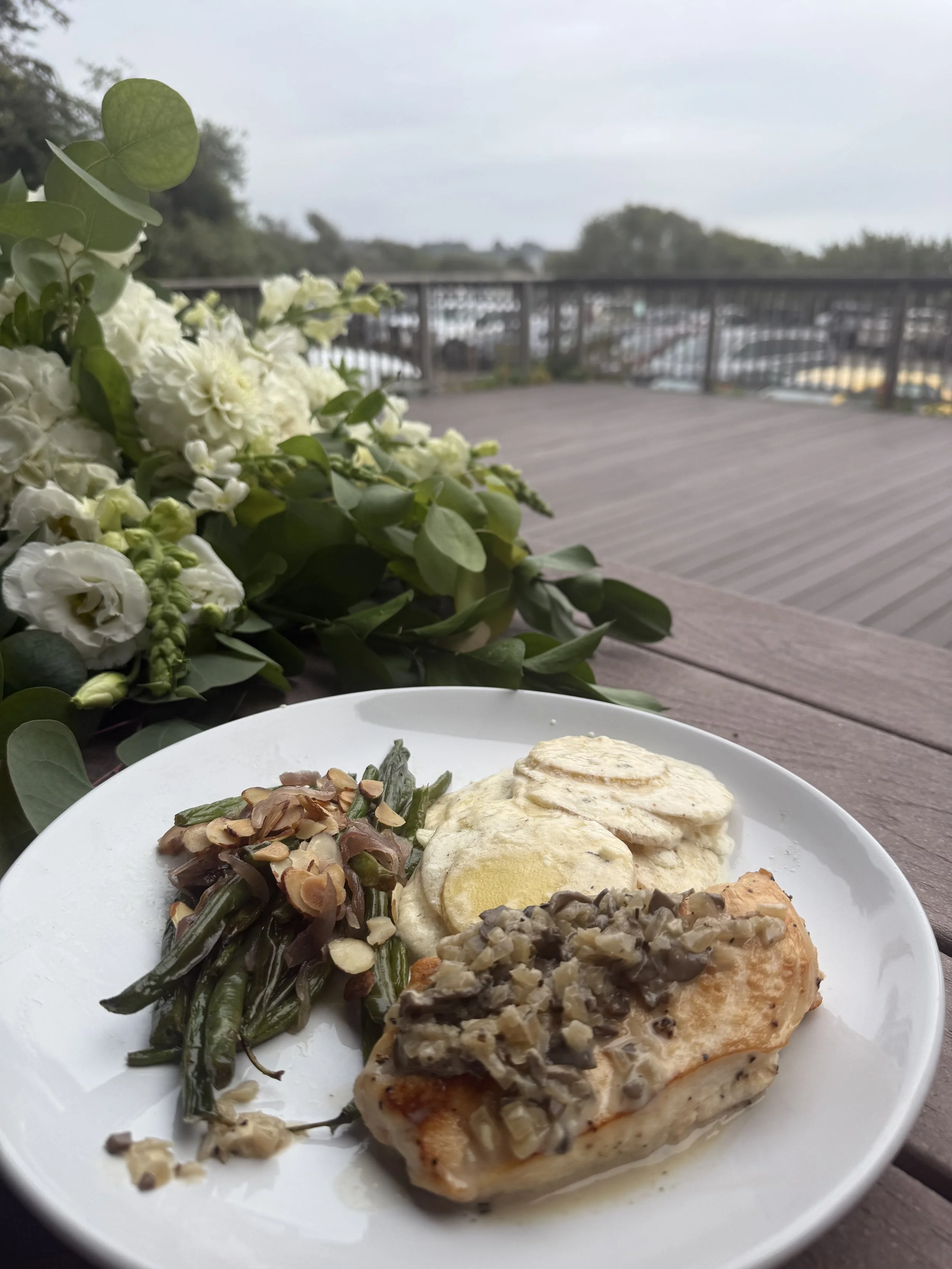 A plate with a serving of chicken topped with a mushroom sauce, green beans with sliced almonds, and mashed potatoes with gravy, placed on a wooden table outdoors, with a bouquet of white flowers nearby and a cloudy sky in the background.