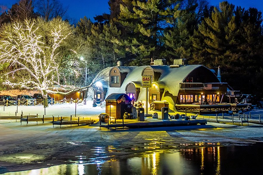 Nighttime scene of a snow-covered building with a curved roof, surrounded by snow and illuminated trees with Christmas lights, reflecting on a frozen surface.