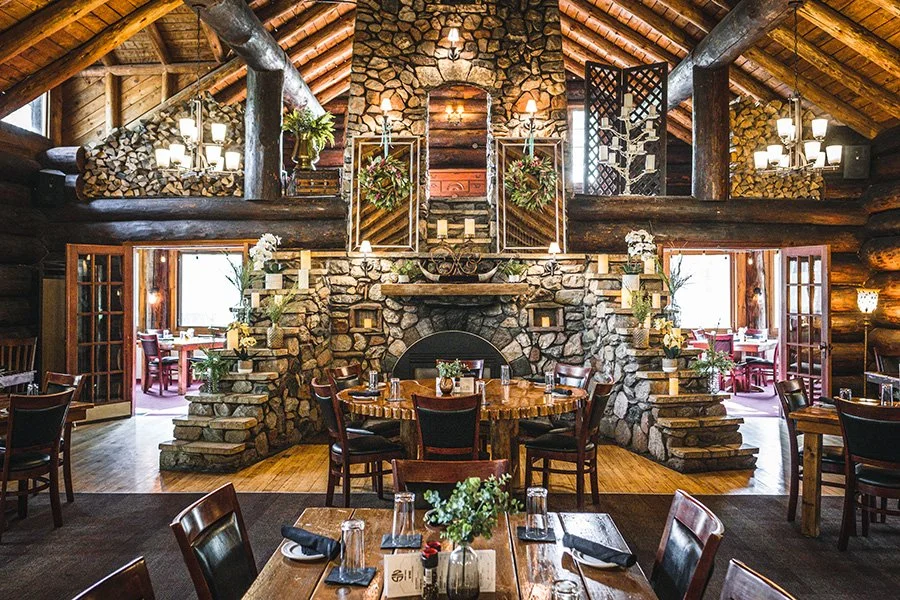 Rustic restaurant interior with stone fireplace, wooden beams, and round wooden tables set with glassware and napkins, decorated with flowers.