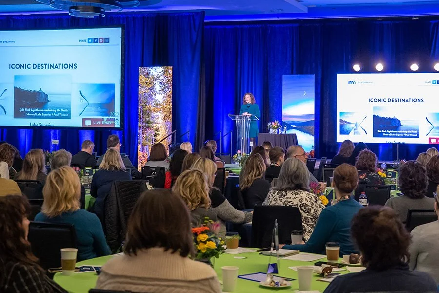 A woman speaking at a conference stage with large screens displaying the presentation titled "Iconic Destinations" and an audience seated at tables with floral centerpieces.