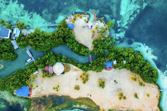 Aerial view of a tropical island with sandy beaches, dense green trees, and colorful buildings near the shoreline, with blue waters surrounding the island.