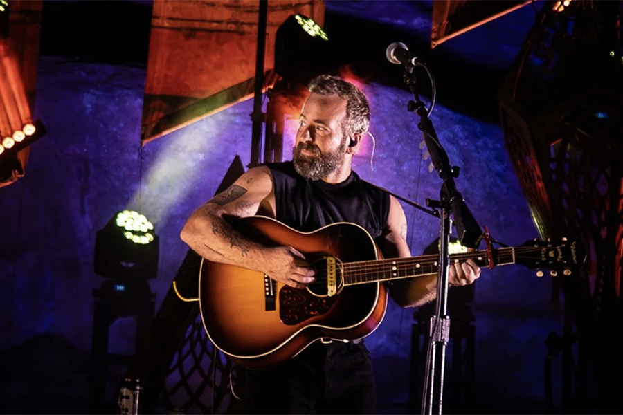 A bearded man with tattoos playing an acoustic guitar on stage, illuminated by colorful stage lights.