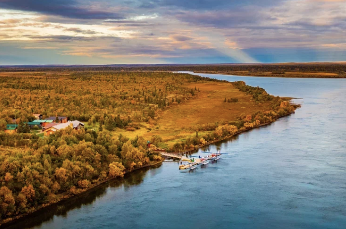 Aerial view of a river with small boats docked, surrounded by dense forest and a few houses, under a colorful cloudy sky.