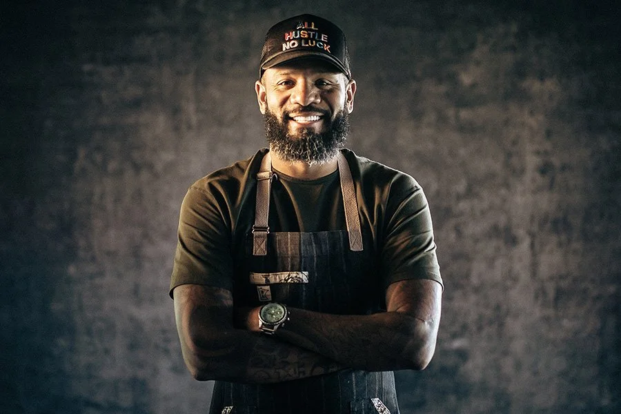 Smiling man with a beard wearing a cap, black apron, and a green shirt, standing with arms crossed against a dark textured background.