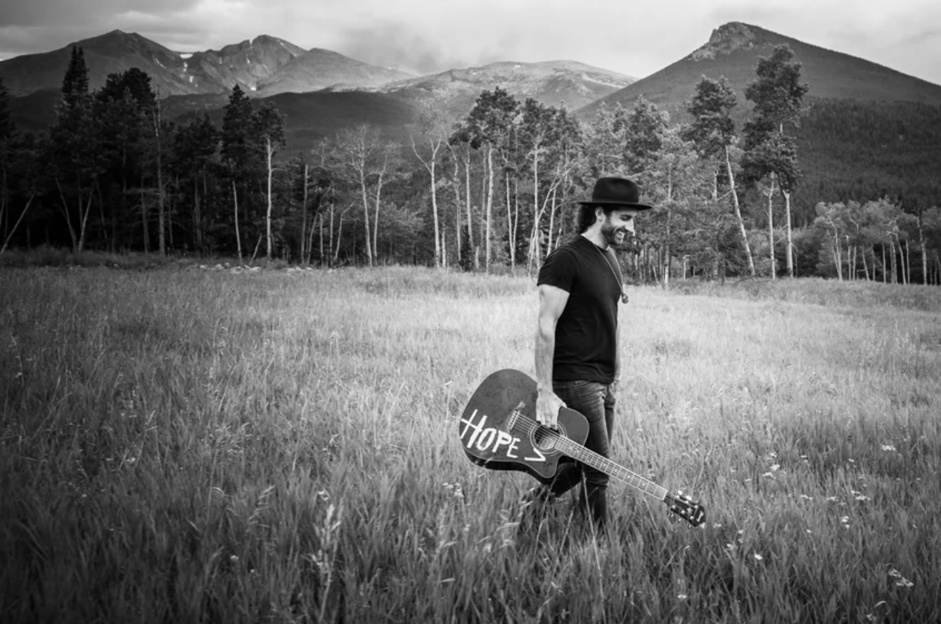 A man with a beard and long hair, wearing a hat and black t-shirt, holding a guitar with the word 'HOPE' written on it, walking through a grassy field with a mountain landscape in the background.
