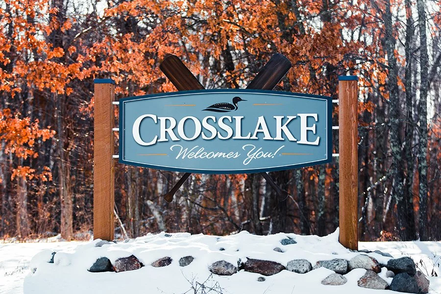 A sign reads 'Crosslake Welcomes You!' with a black duck silhouette, flanked by two oars, in a snowy outdoor setting with autumn trees in the background.