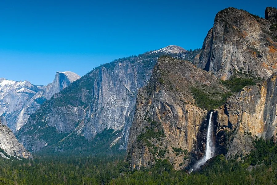 Scenic view of a mountainous landscape with a waterfall cascading down a rock face, dense green forest at the base, and snow-capped peaks in the background under a clear blue sky.