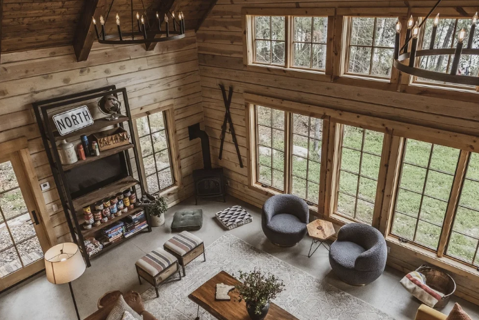 Interior of a cozy wooden cabin living room with large windows, a black wood stove, two gray chairs, a bookcase, a wooden coffee table, and rustic decor.