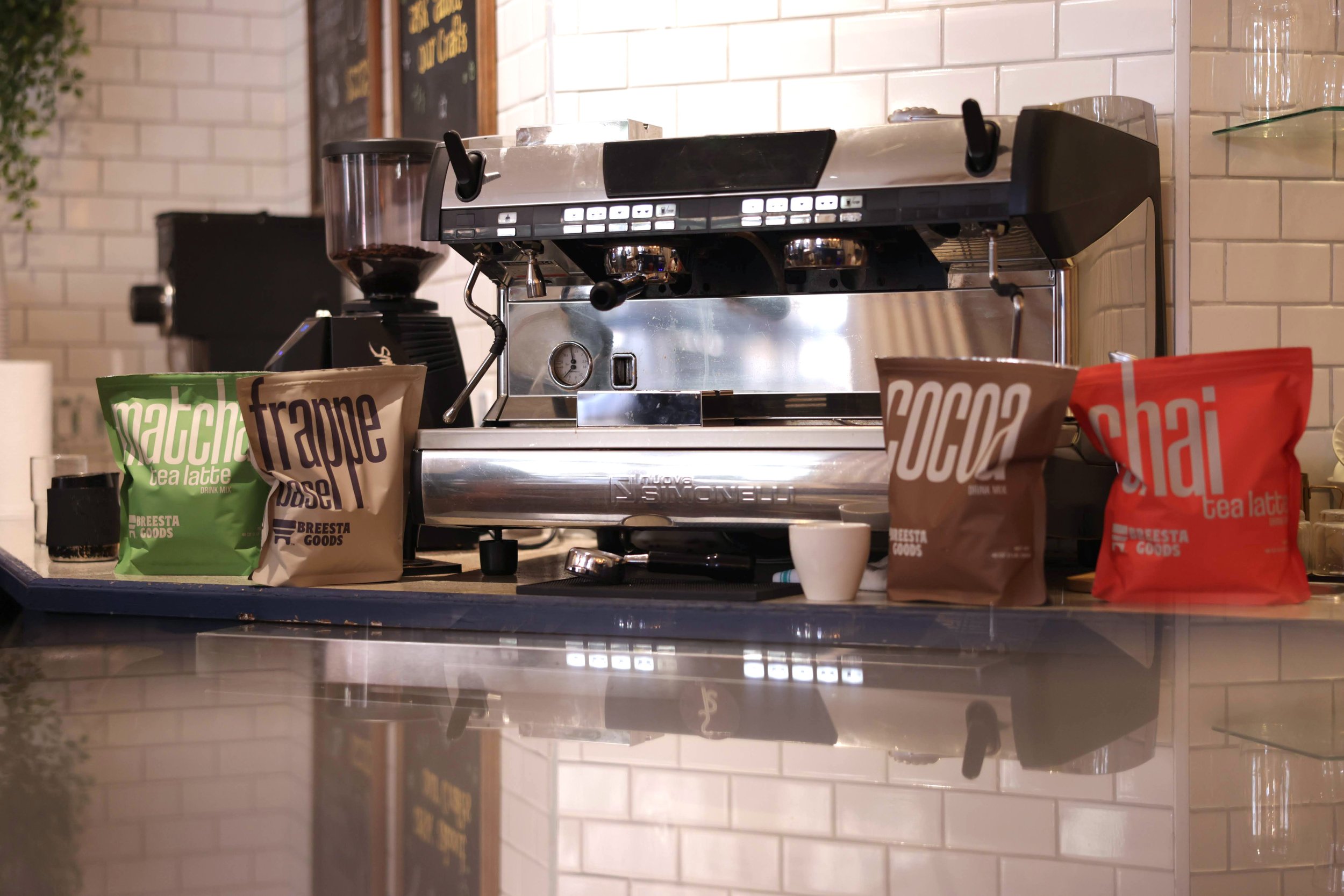 A stainless steel espresso machine on a black countertop, with bright colored Breesta Goods powder bags and a small white cup in front, in a cafe setting with white tiled walls.