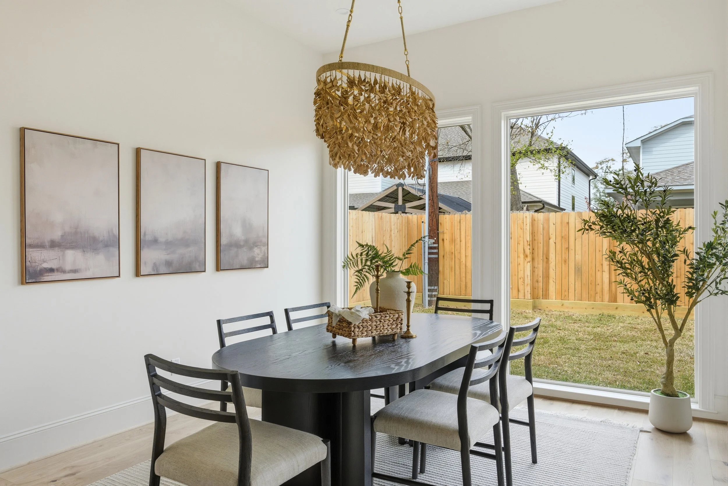Dining room with a black table, six chairs, a large window showing a backyard with a wooden fence, and a potted tree inside.