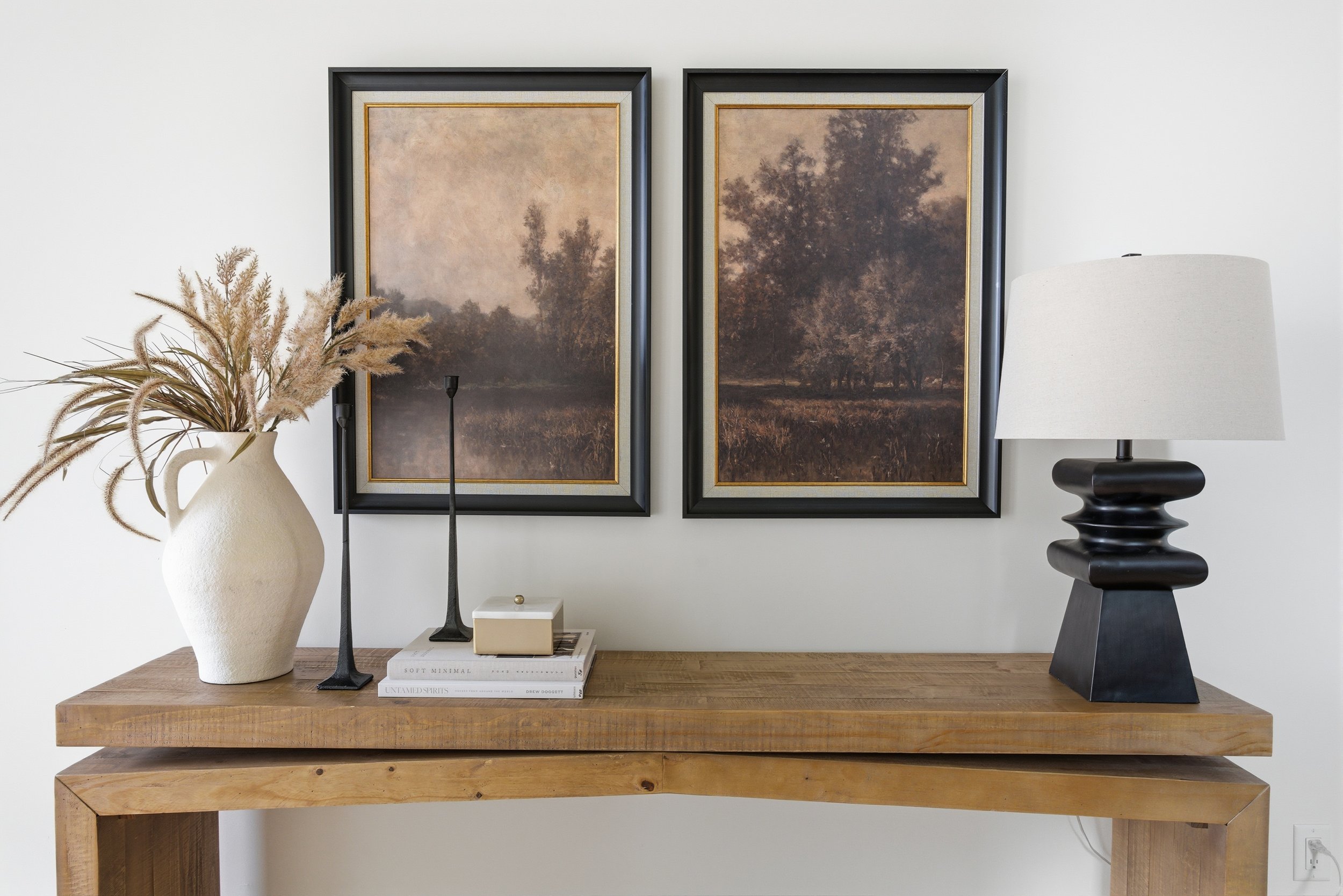 Decorative wooden console table with beige vase filled with pampas grass, two black candlesticks, a small beige box, framed landscape paintings of trees and water, and a black table lamp with a white shade against a white wall.