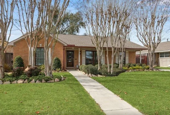 A brick house with a gray roof, front porch, and large windows, surrounded by a well-maintained lawn with trees and shrubs.