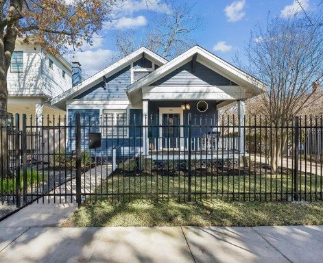 A blue house with white trim, a front porch, and a black metal fence in front, with trees and a sidewalk in the neighborhood.