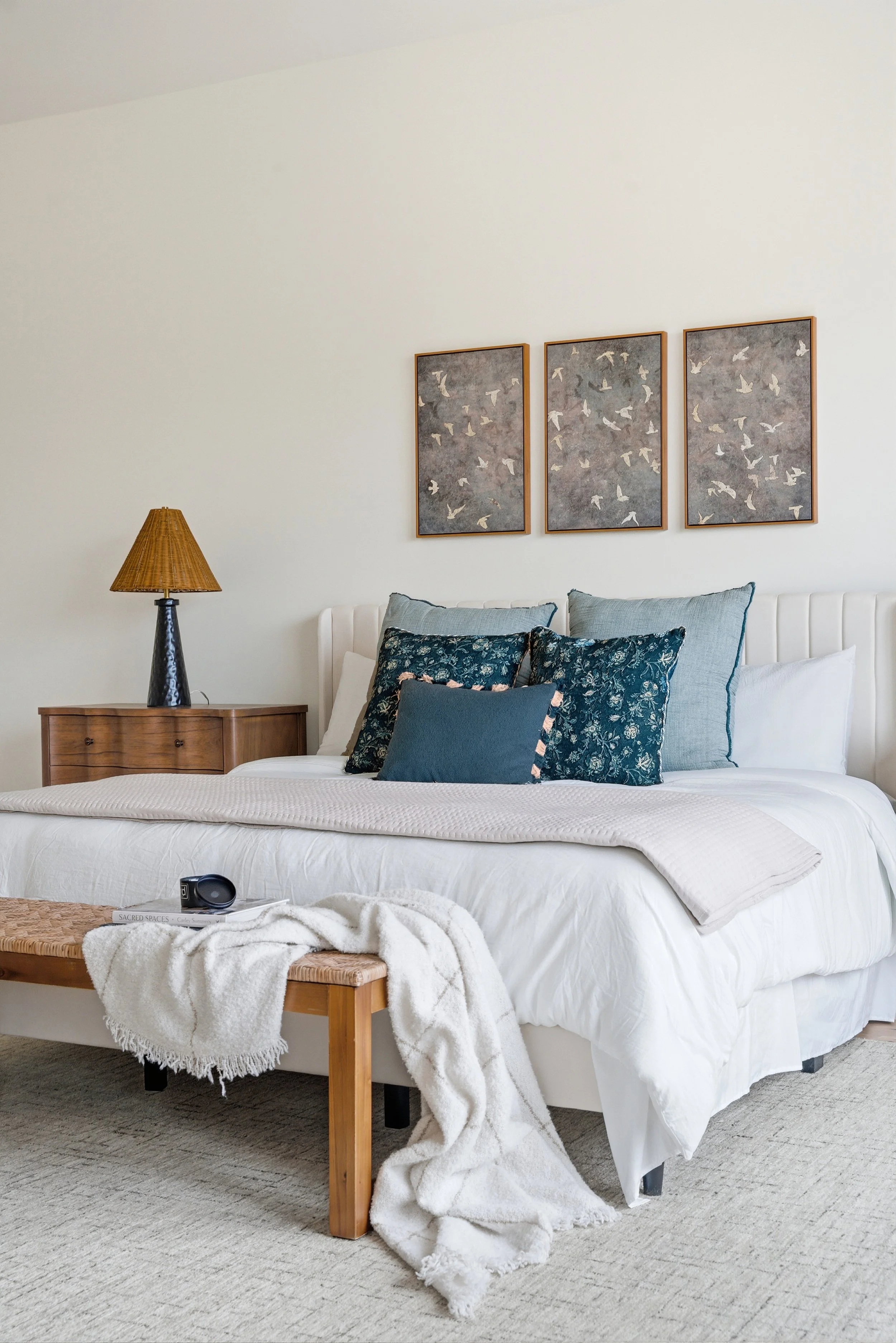 A neatly made bed with white bedding, multiple decorative pillows in shades of blue, a wooden bench with a white blanket draped over it, a bedside lamp on a wooden nightstand, and three framed art pieces of birds on the wall behind the bed.