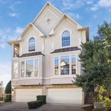 A large, multi-story residential house with beige exterior, white trim, and multiple large windows. The house has a sloped roof, a small visible balcony, and a three-car garage. There are trees and bushes in front and to the side.