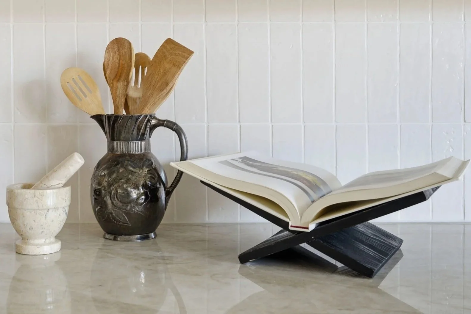 A metal pitcher holding wooden cooking utensils, a small white mortar and pestle, and an open book on a black stand, on a marble countertop against a white tiled wall.